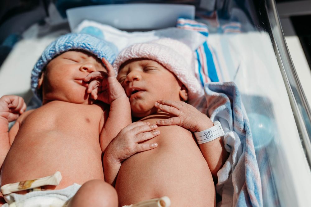 Two newborn twins in blue and pink hats lying together in a hospital bassinet after birth.