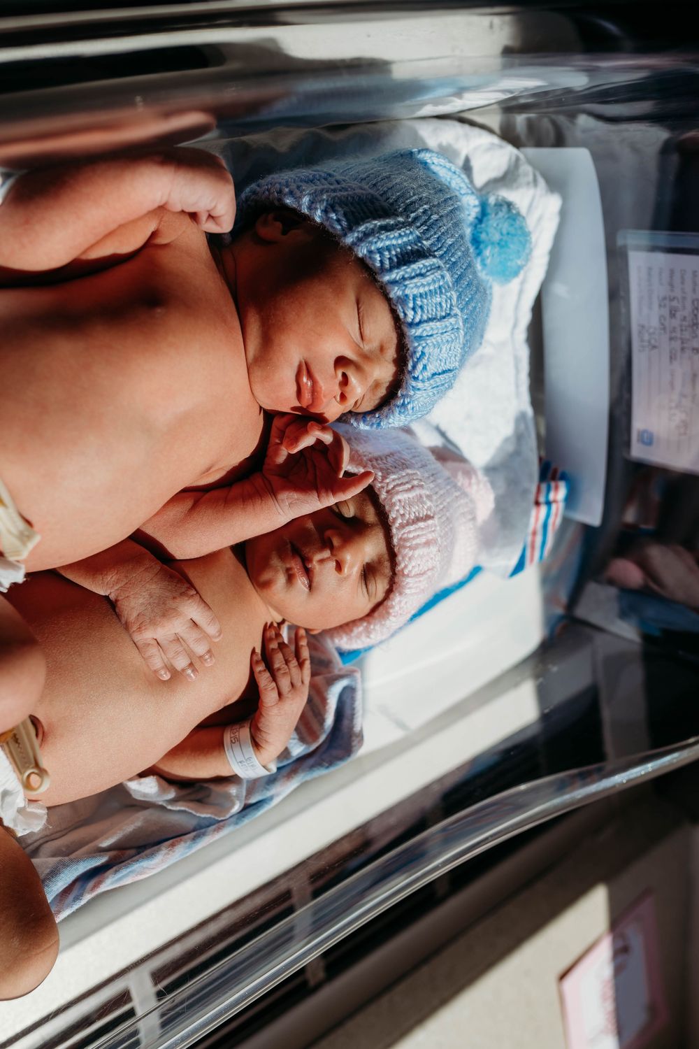 Two newborn twins in knitted hats resting together in a hospital bassinet.