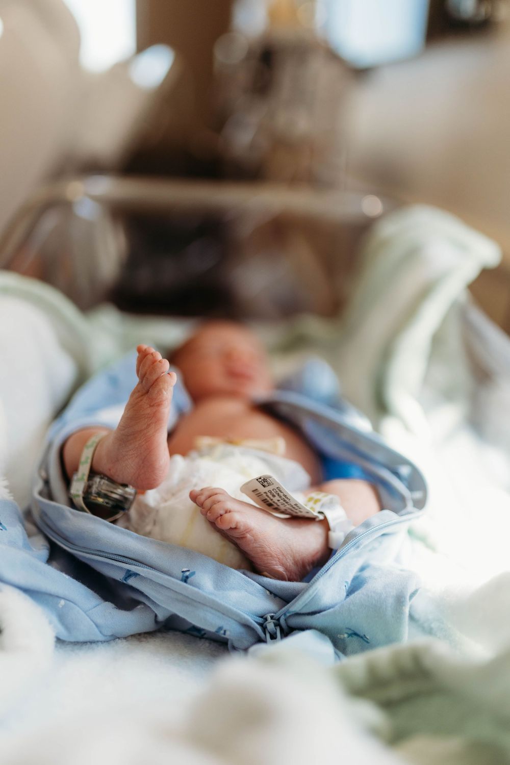 Newborn baby resting in hospital incubator wrapped in blue blanket with medical ID bracelet on ankle.