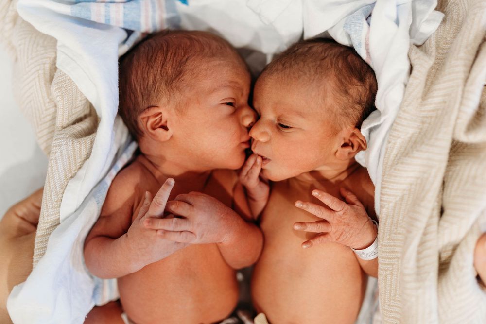 Two newborn twins lying face-to-face sharing a sweet kiss, wrapped in soft white blankets.