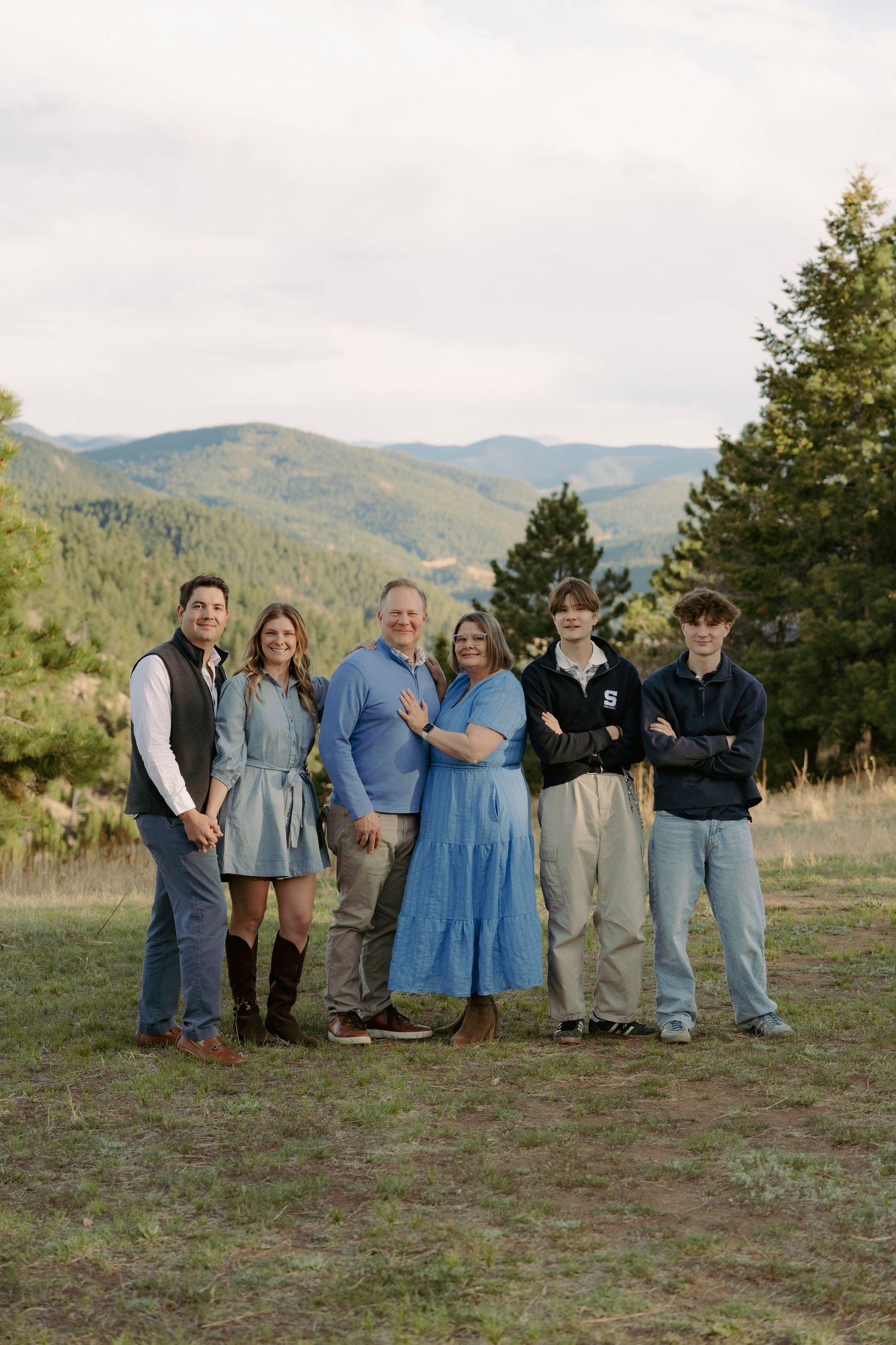 Family of six posing outdoors in blue outfits with scenic mountain and forest backdrop.