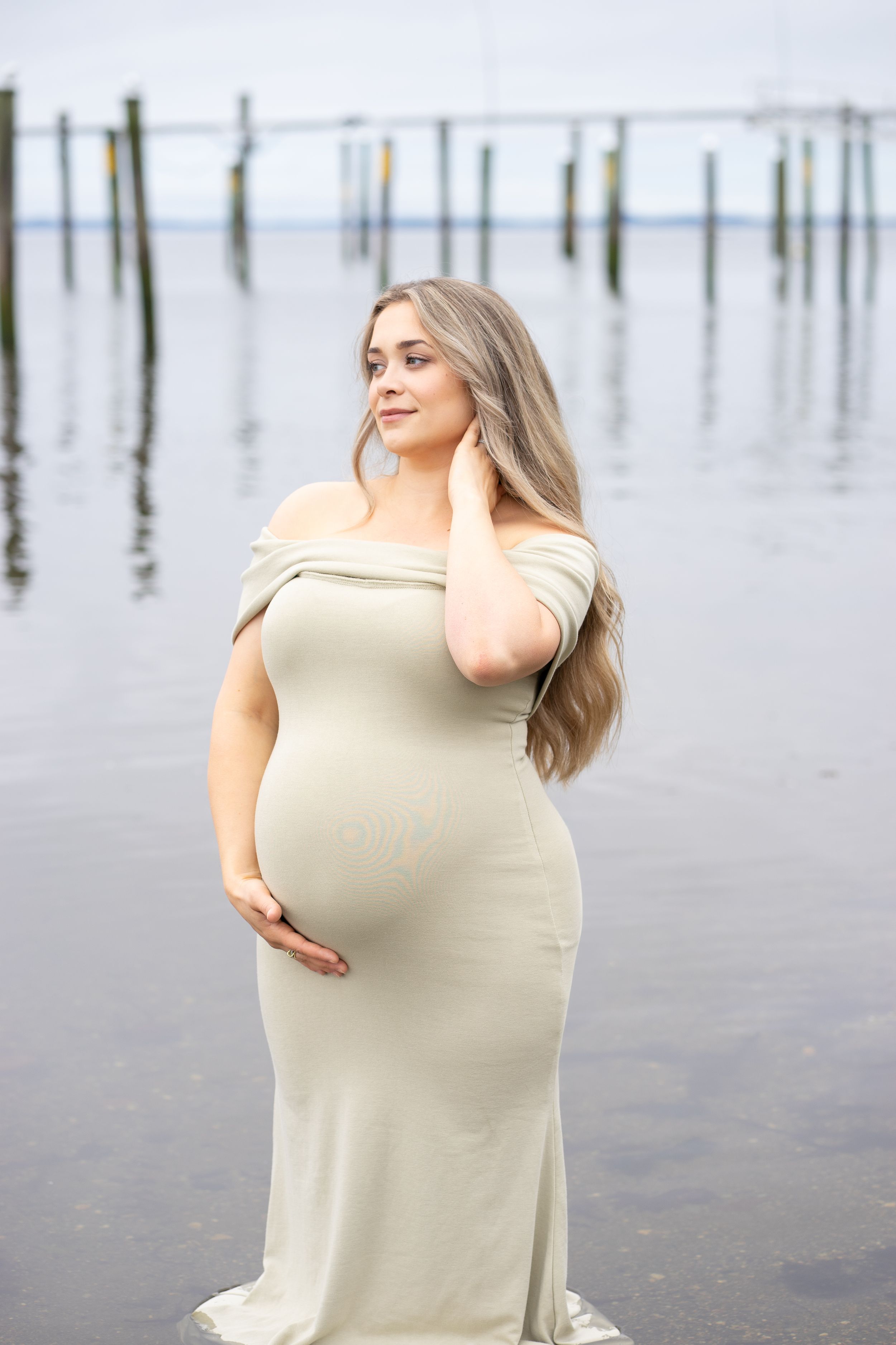 Pregnant woman in sage green off-shoulder dress touching neck while standing in calm water.