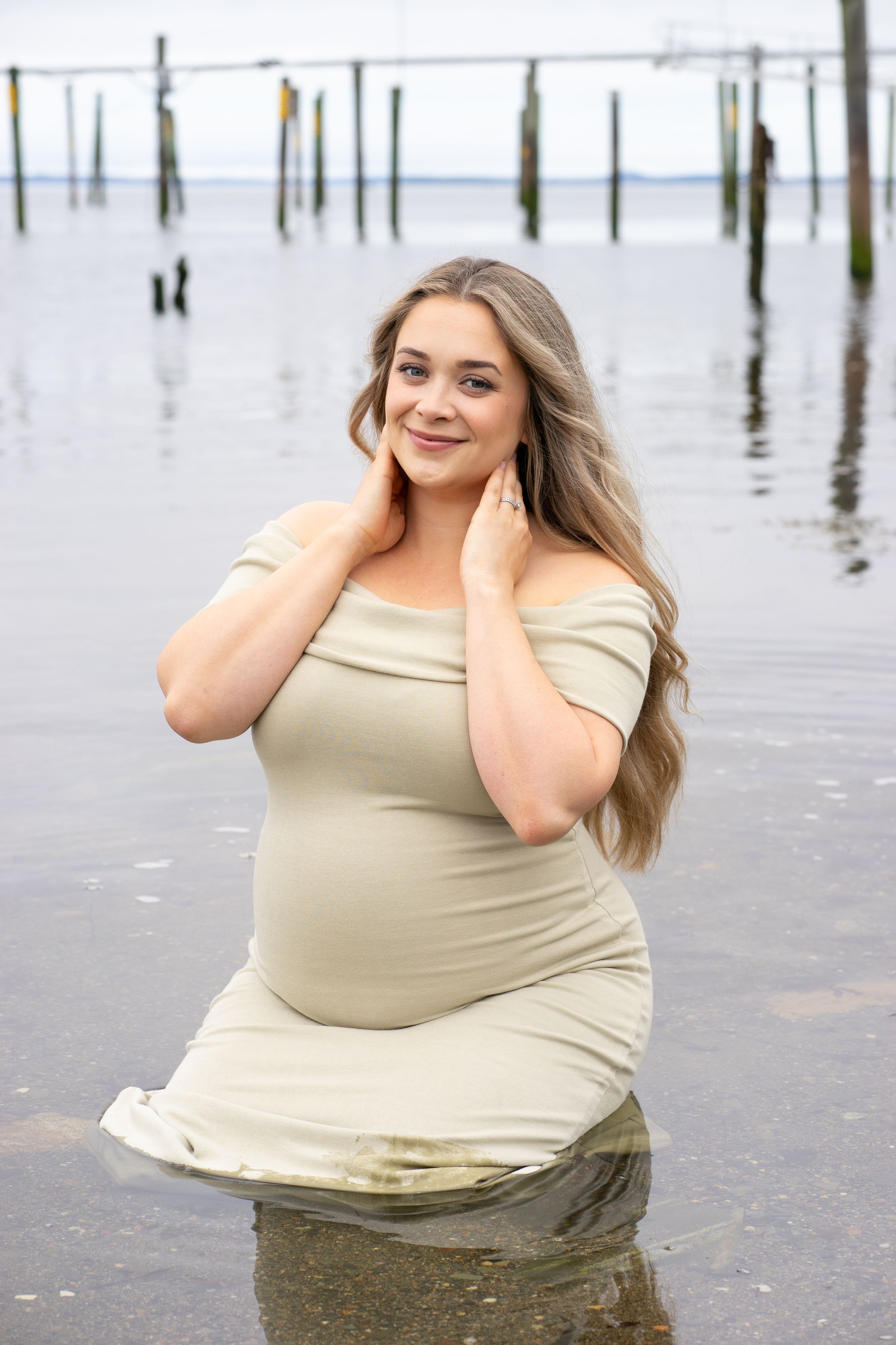 Smiling pregnant woman in sage green dress posing in shallow water with old pier behind.