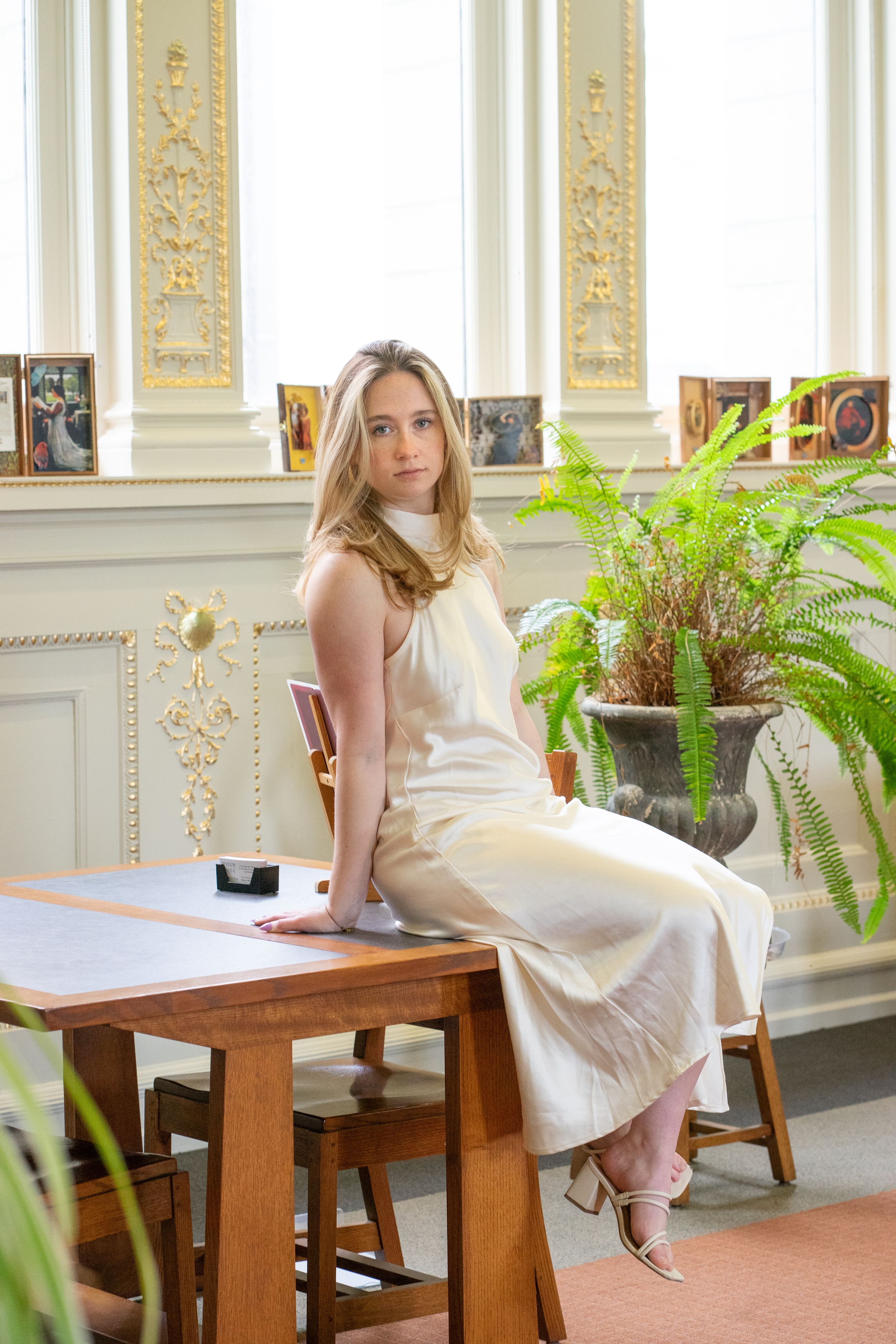 Blonde woman in white sleeveless dress sitting on wooden table in ornate sunlit room.