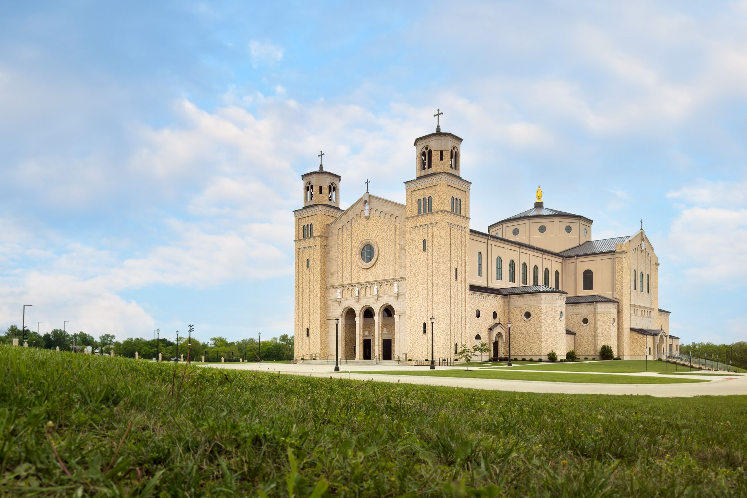 Large ornate Catholic basilica with twin towers and golden dome under blue sky surrounded by green lawns.