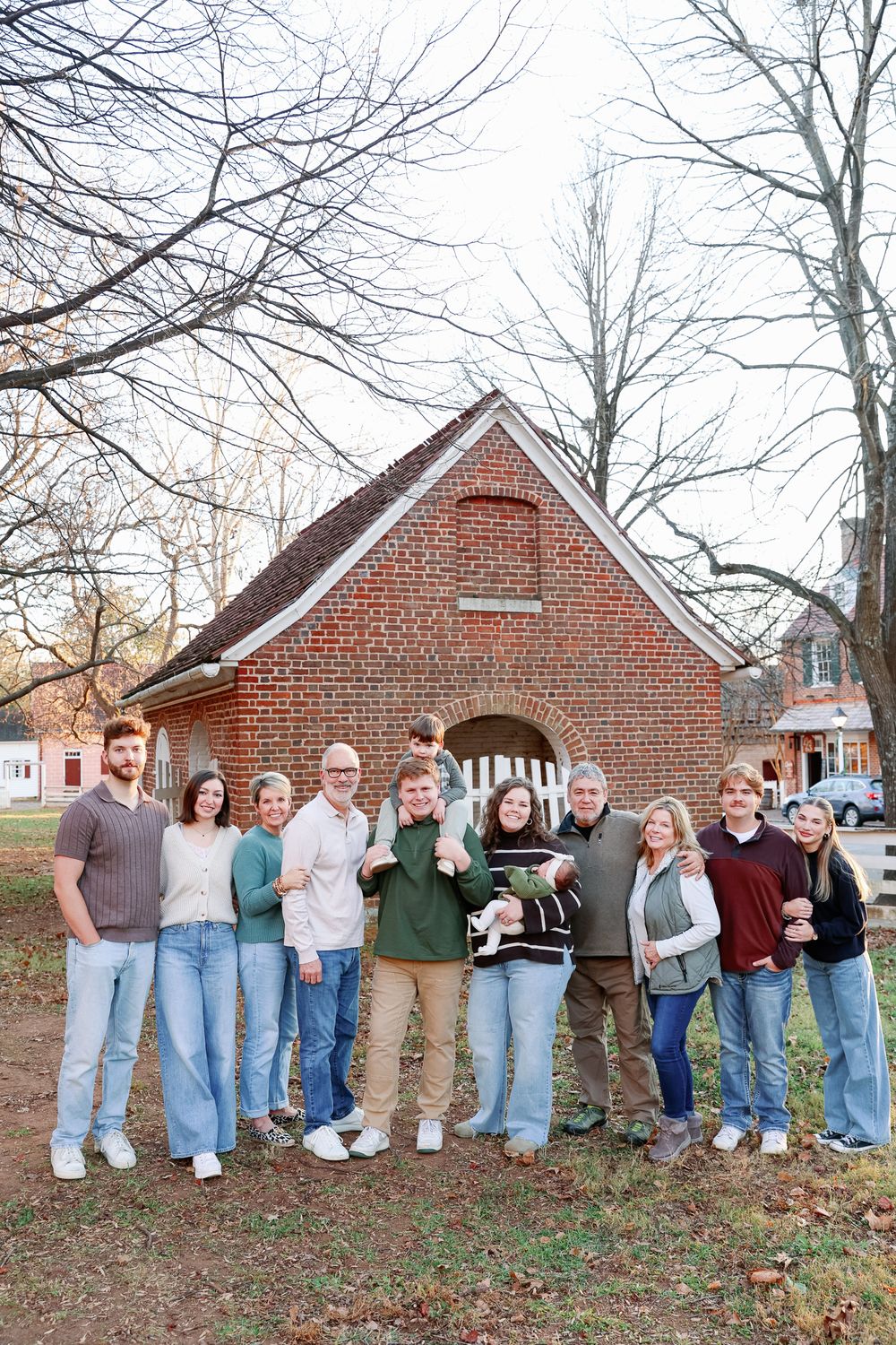 A large family group poses together outdoors in front of a rustic brick building during autumn.
