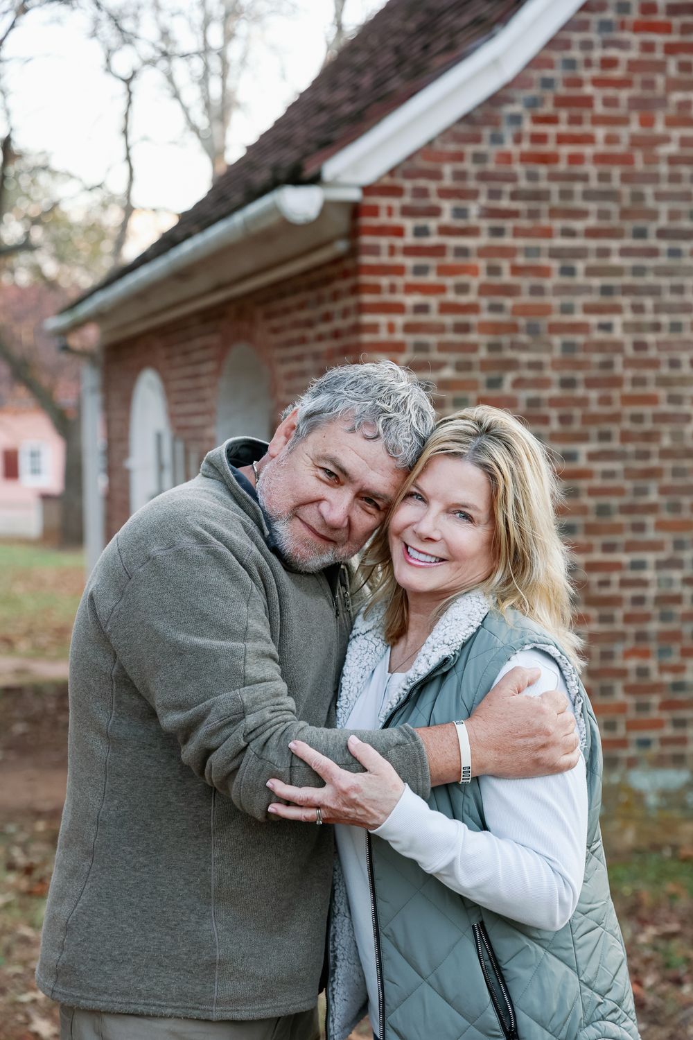 Middle-aged couple embracing and smiling outdoors near a brick building in autumn.
