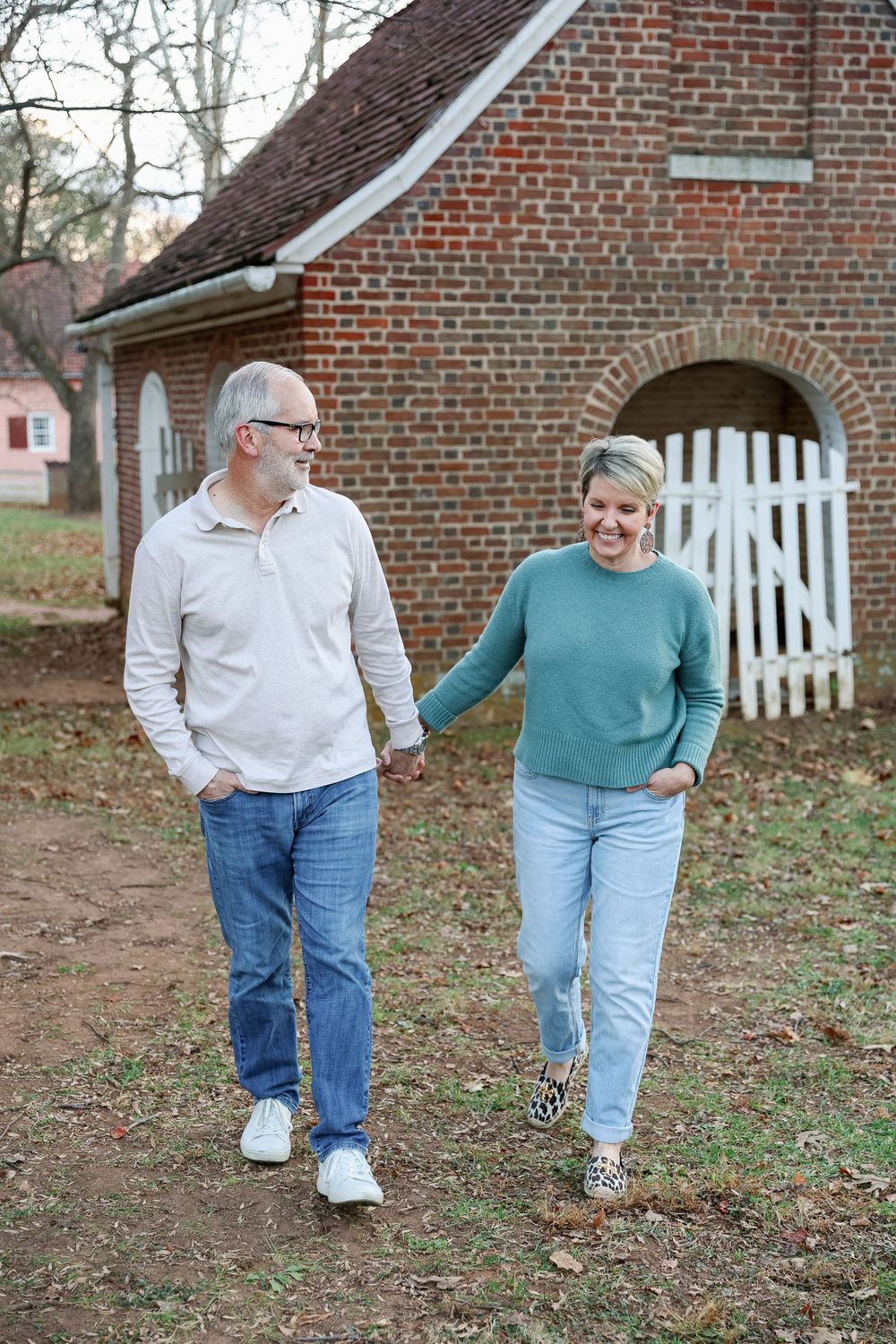 Smiling middle-aged couple holding hands and walking outdoors near a rustic brick building with a white gate.
