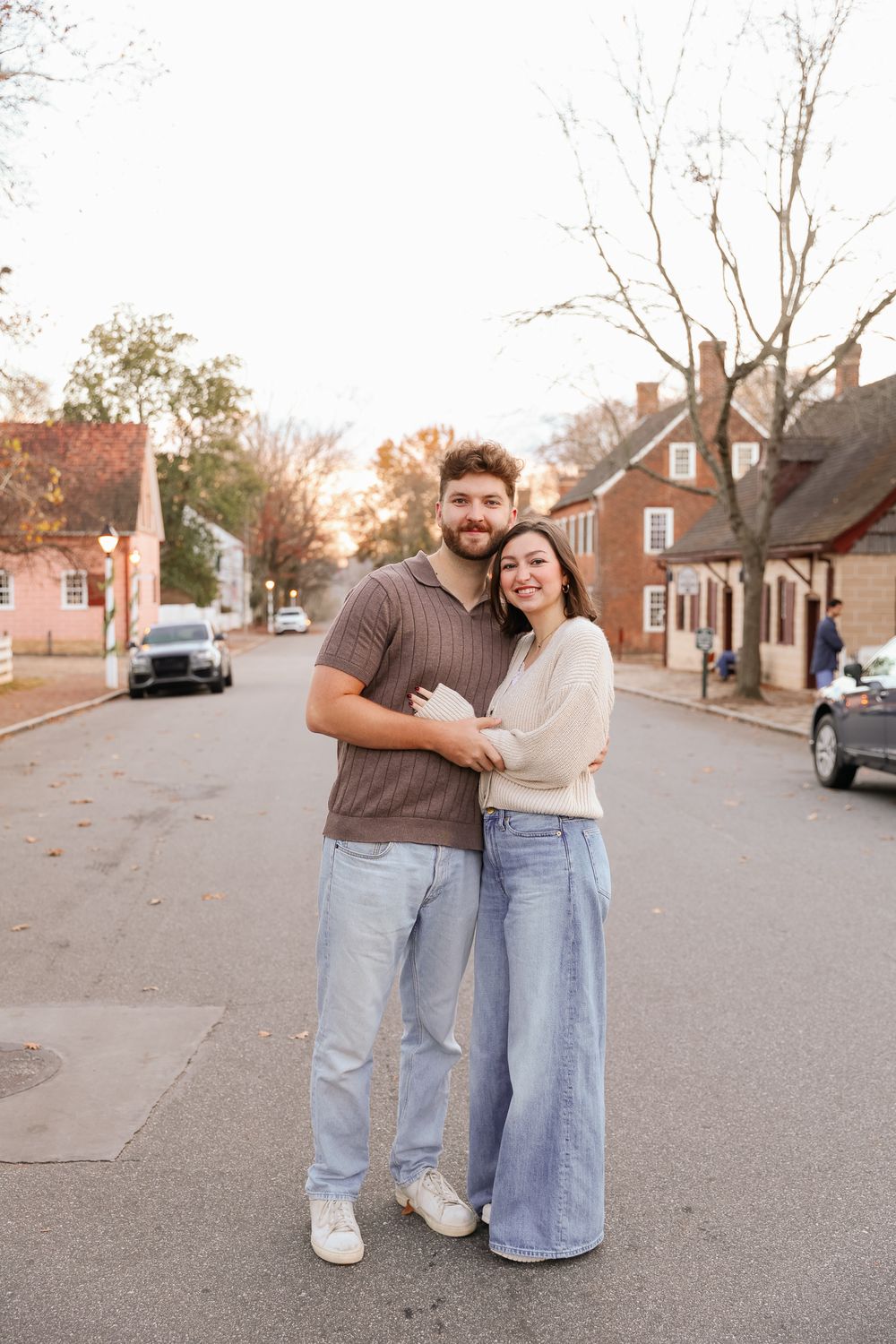 A smiling couple embraces on a quiet historic street at golden hour, wearing casual jeans and neutral tops.