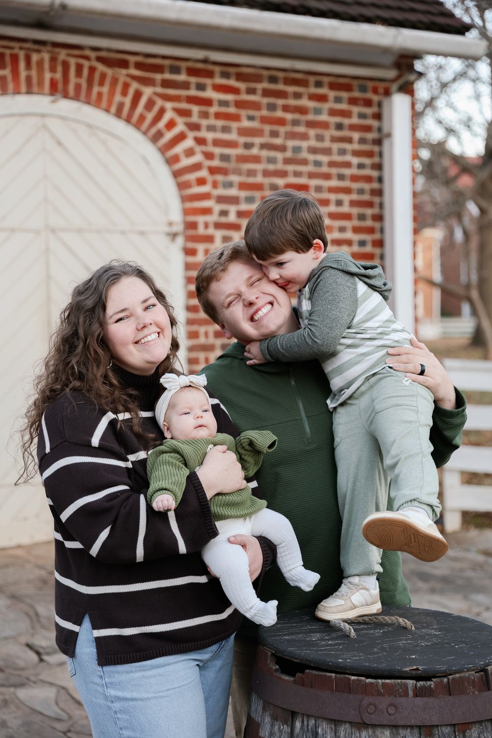 Happy family of four posing outdoors near brick building, parents smiling with toddler and baby in green outfits.