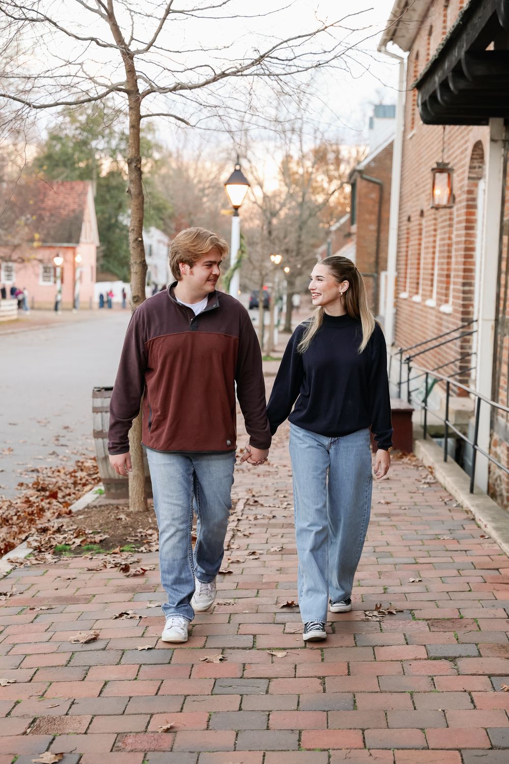 Couple holding hands and smiling while walking on a brick sidewalk in a charming town during autumn.