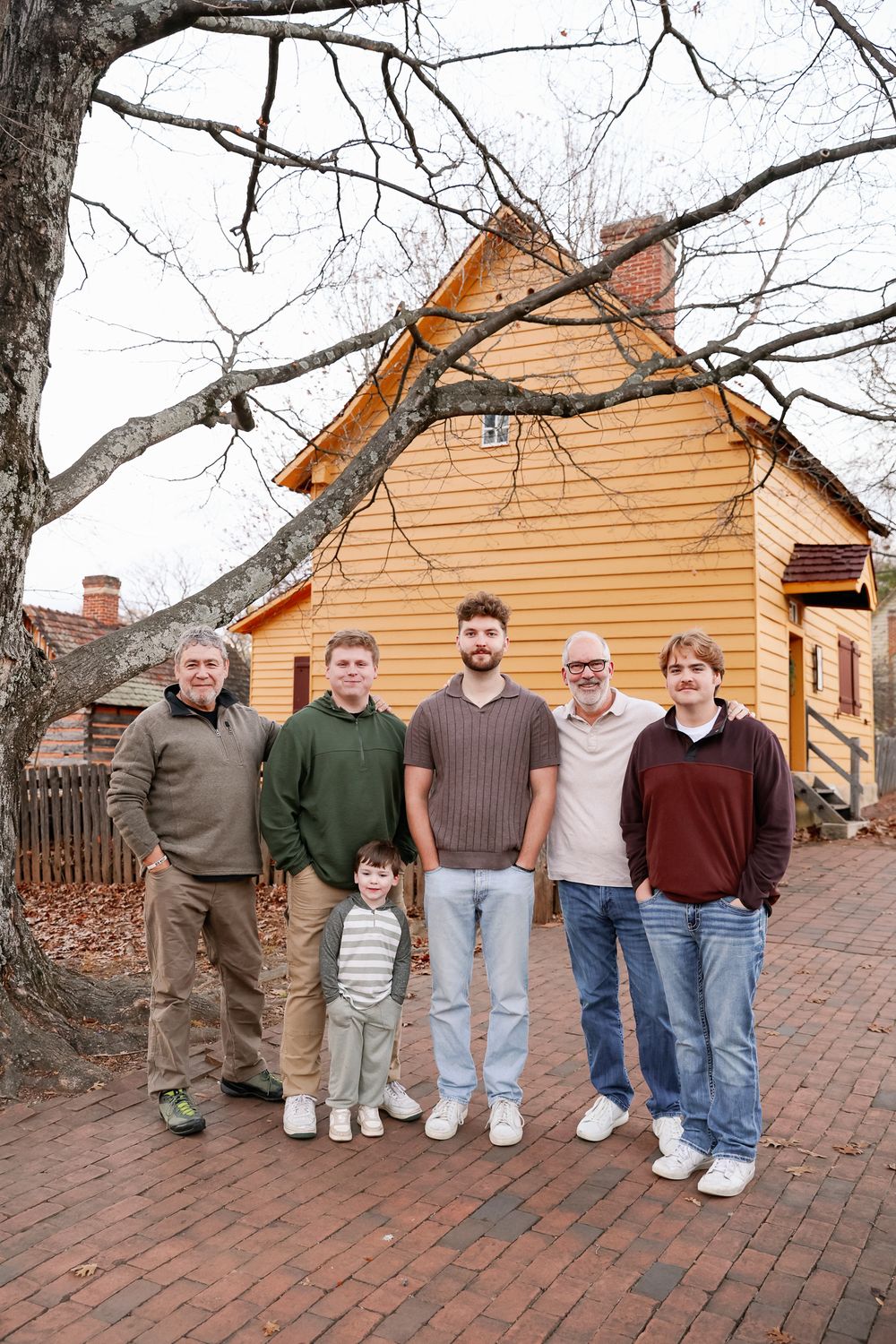 Multi-generational family of six males posing outdoors in front of a yellow historic building on a brick pathway.