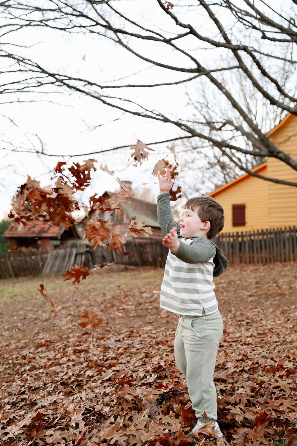 Young boy joyfully tossing autumn leaves in a yard with bare trees and rustic buildings in the background.