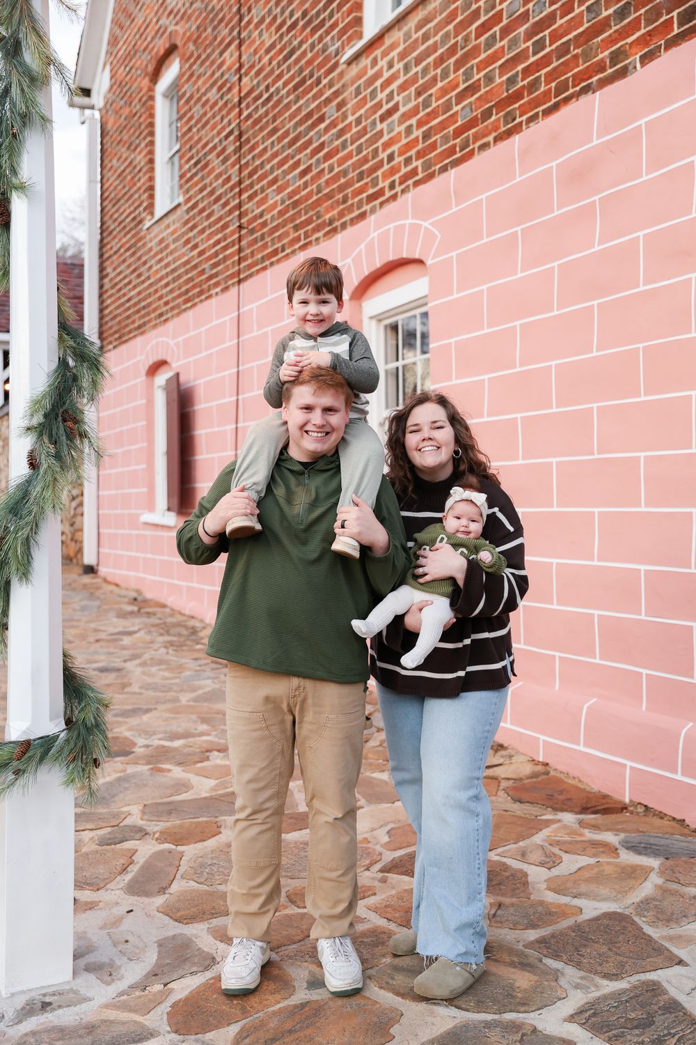 Happy family of four posing outdoors near a pink brick wall, parents holding two young children on a stone patio.