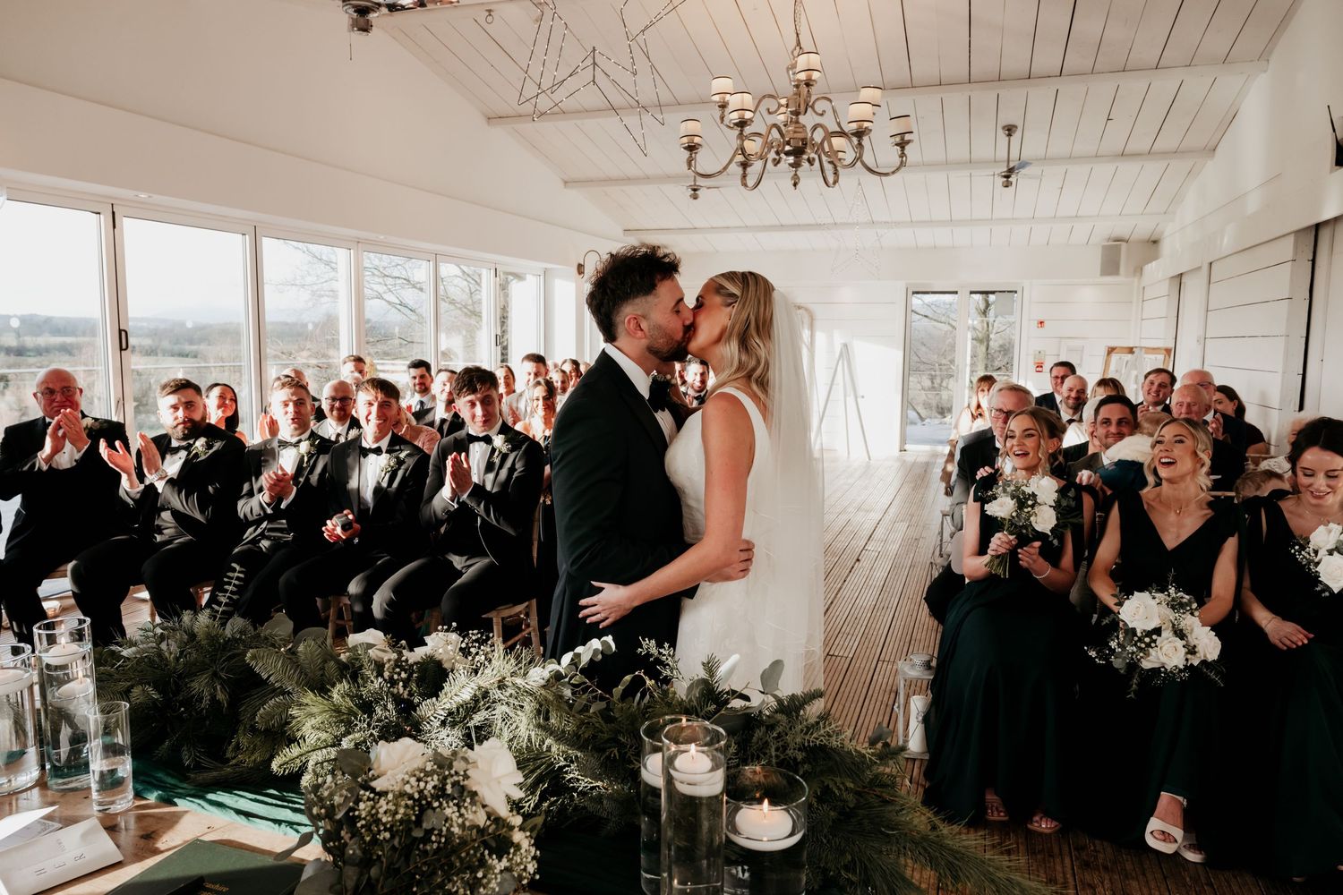 Bride and groom share first kiss at indoor wedding ceremony surrounded by guests in formal attire and floral arrangements.