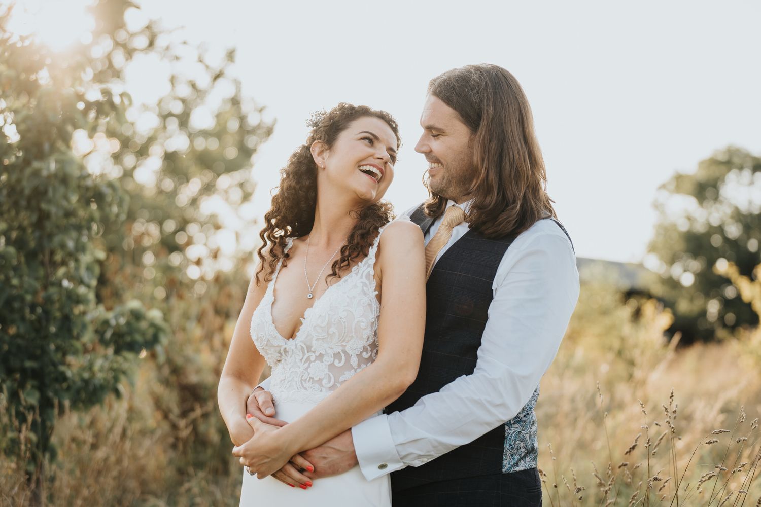 A joyful bride and groom embrace outdoors in golden summer sunlight, laughing together in a field during their wedding day at Oxleaze Barn
