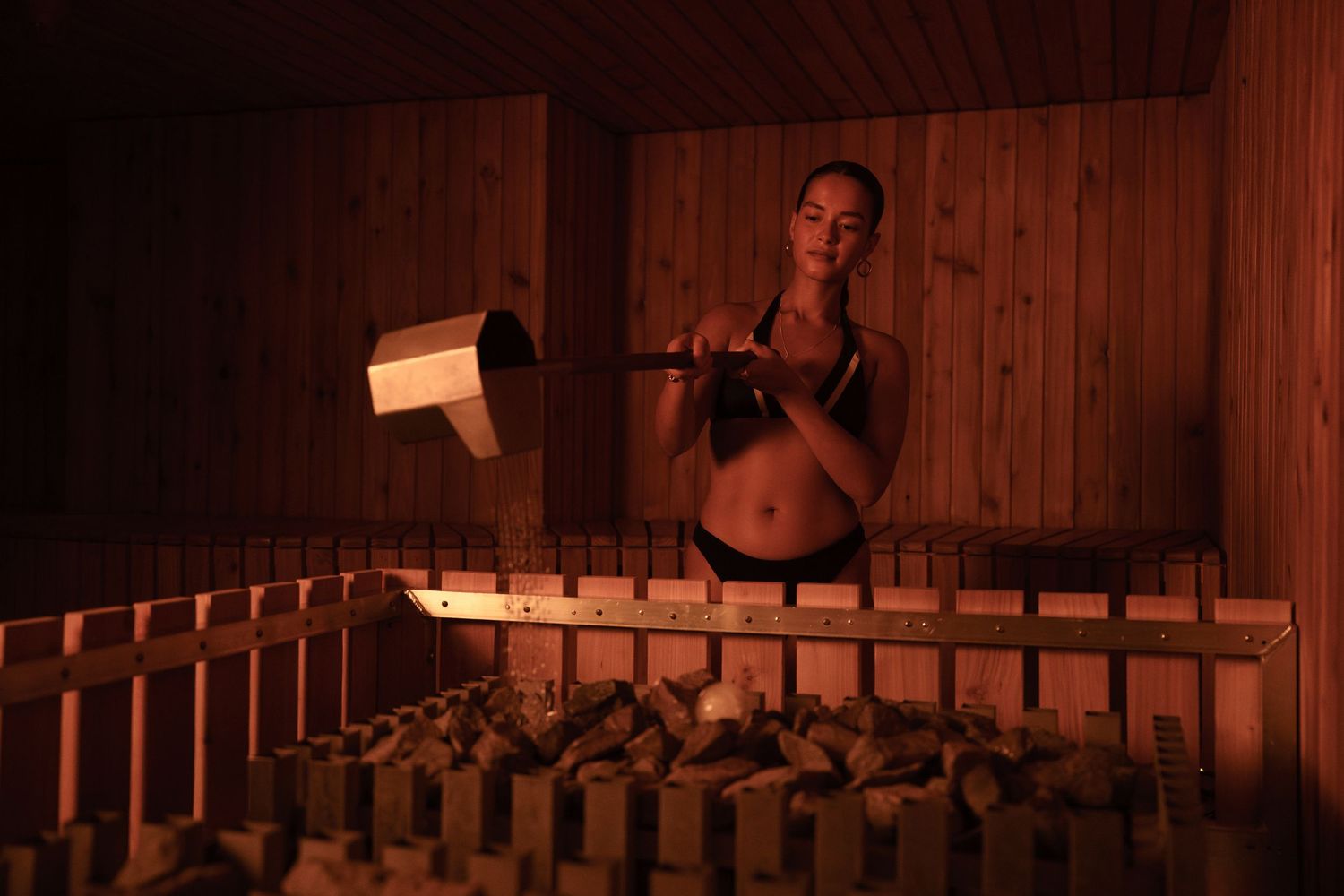 Woman in bikini top pouring water on sauna rocks with wooden ladle in a dimly lit wooden sauna.