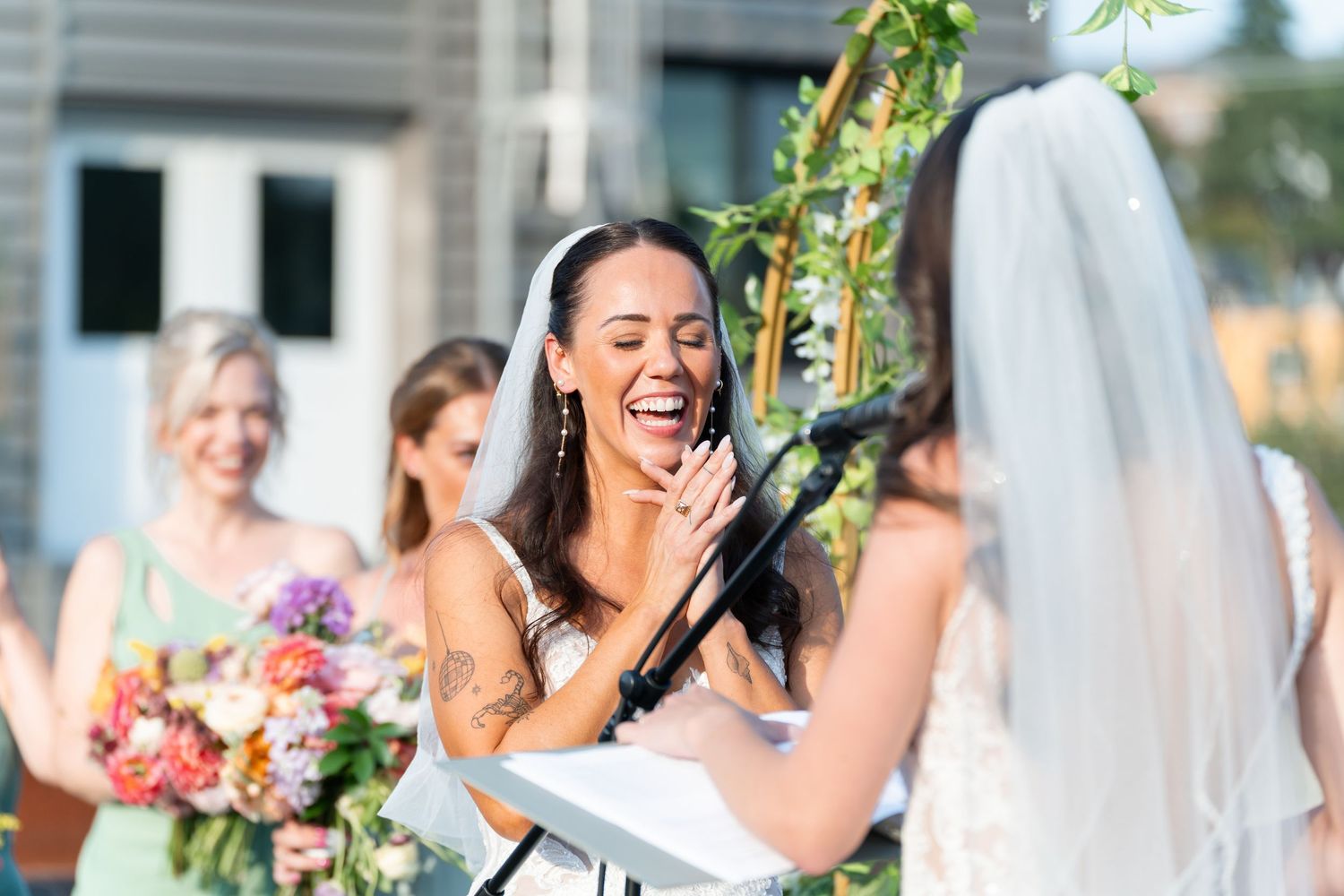 Bride and groom laughing joyfully at outdoor wedding reception dinner table with floral arch and champagne.