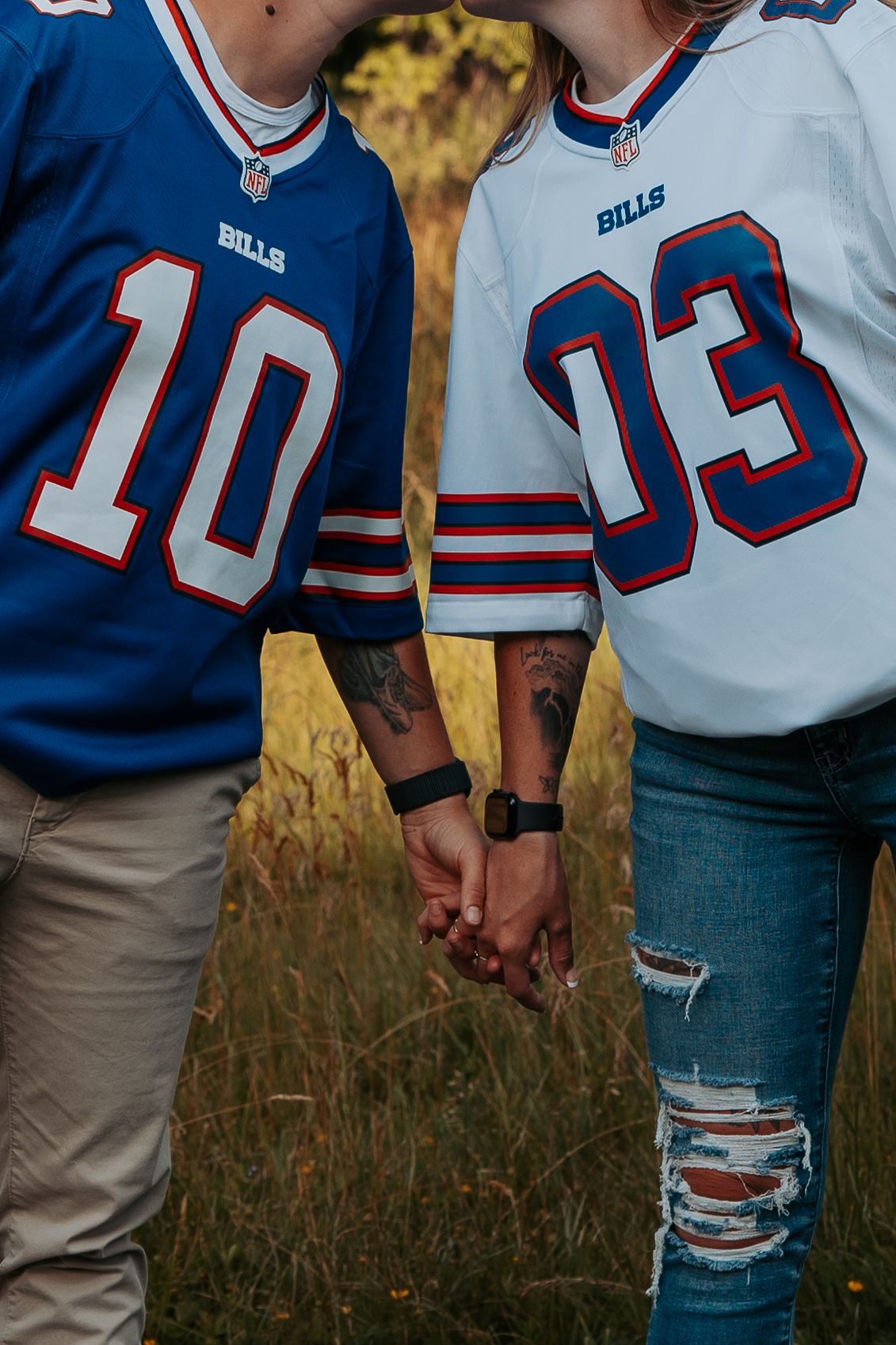 Couple holding hands in Buffalo Bills jerseys numbered 10 and 03, showing tattoos and ripped jeans outdoors.