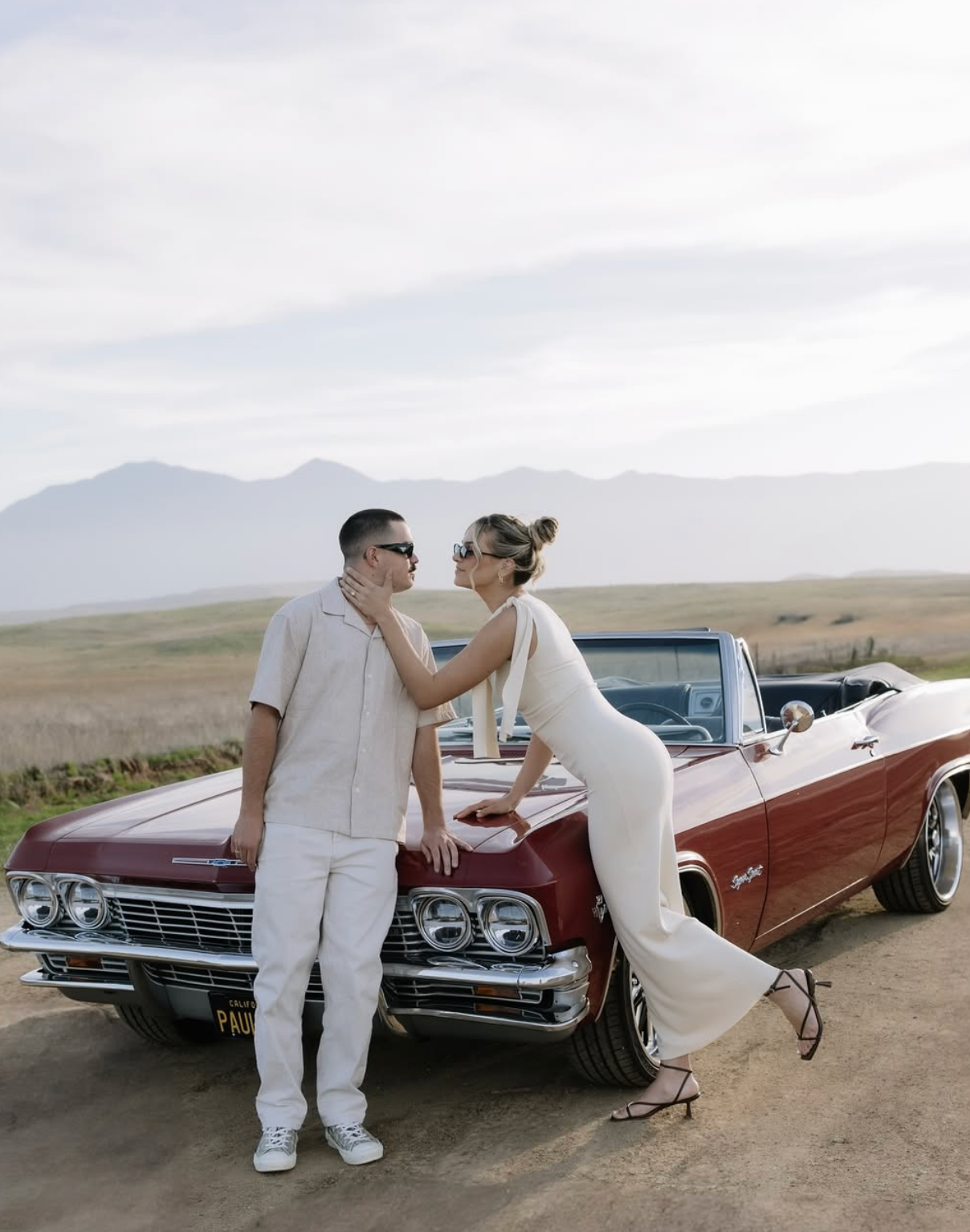 Couple in white outfits leaning on a vintage red convertible with mountain backdrop.
