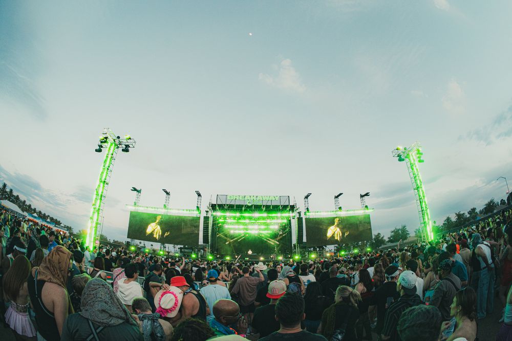 Large outdoor music festival crowd gathered before a bright green-lit stage at dusk, captured with fisheye lens.
