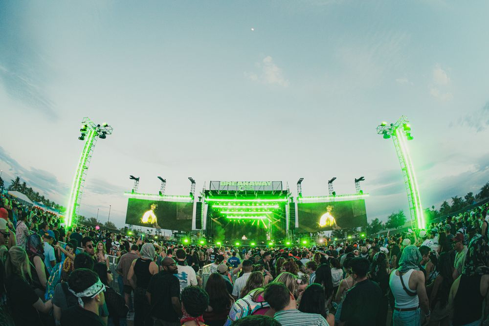 Large outdoor music festival crowd enjoys a concert with bright green neon stage lights at dusk.