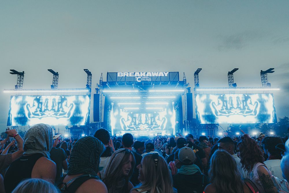 Breakaway music festival crowd enjoys a vibrant light show on an illuminated stage at dusk.