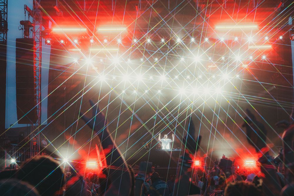 Crowd at an outdoor music festival with dramatic red and white laser light show illuminating the stage at night.