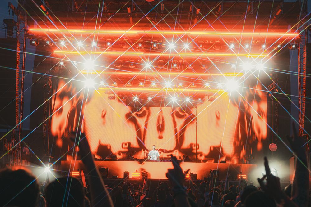 Crowd at outdoor music festival with vibrant orange stage lights and laser beams illuminating the night sky.