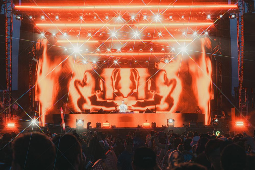 Crowd at an outdoor music festival with a vibrant red-lit stage and dramatic shadow projections.