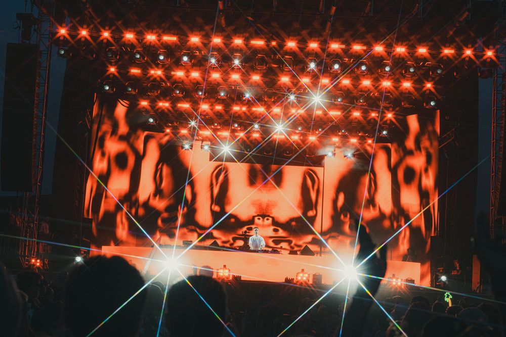 DJ performing on a vibrant festival stage with orange lights, laser beams, and dynamic visual projections at night.