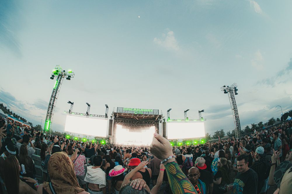 Large outdoor music festival crowd faces a bright stage with green lights under a cloudy sky.
