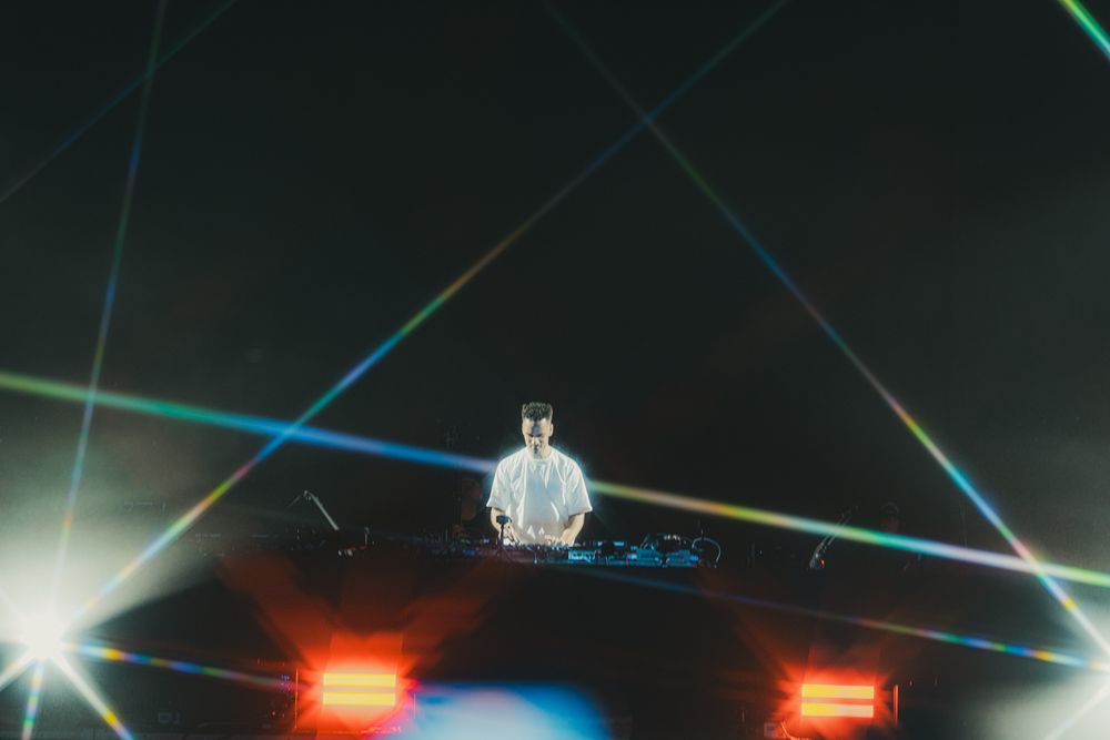 DJ performing on stage surrounded by colorful laser lights in a dark concert venue.
