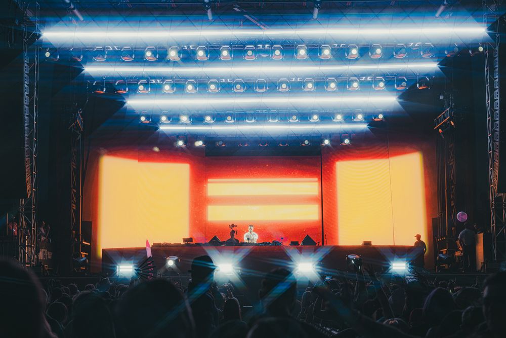 Concert stage with vibrant orange screens, bright LED lights, and a crowd of fans holding up phones.