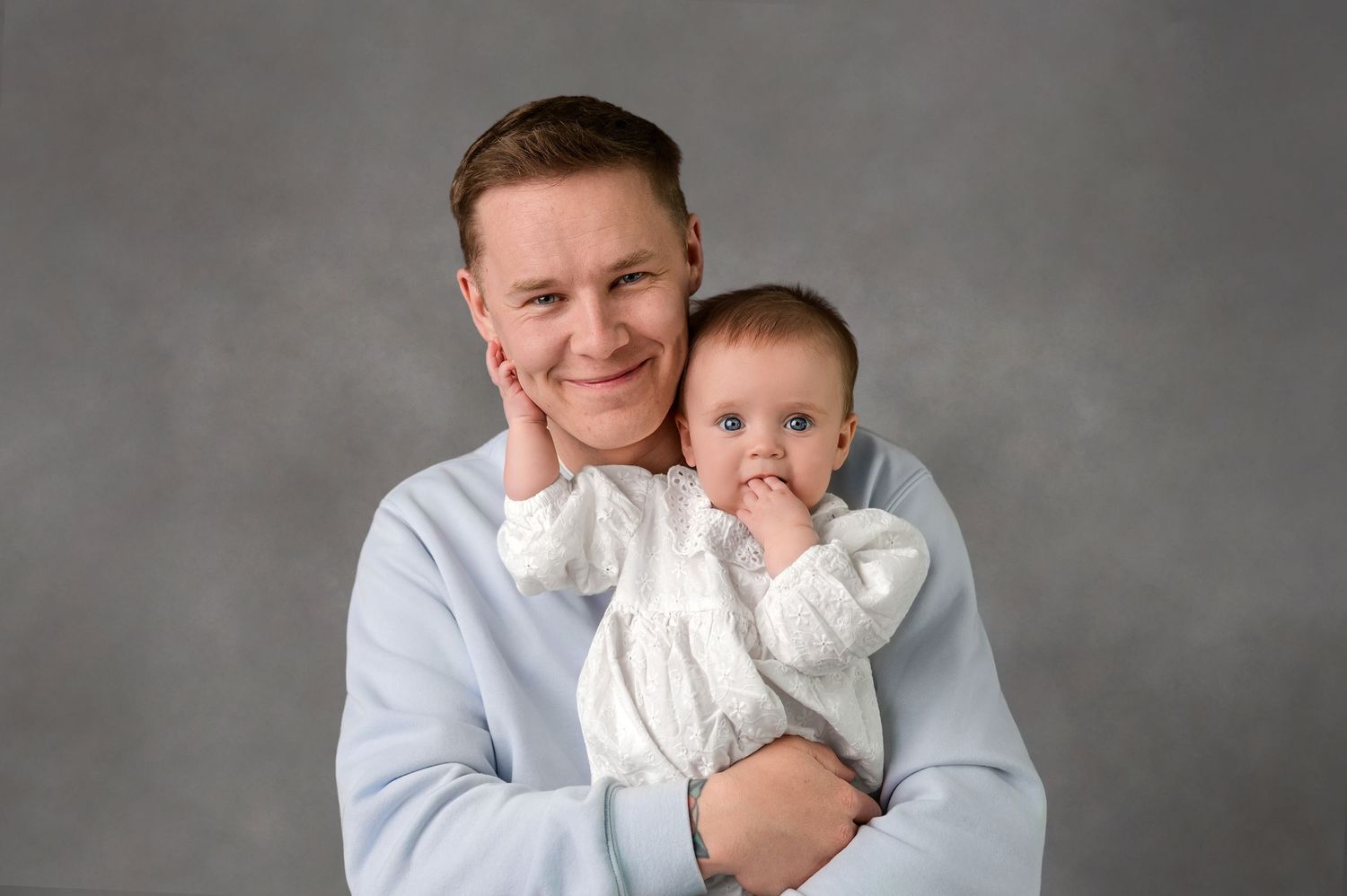 Smiling man in light blue sweater holding a cute baby in white outfit against gray background.