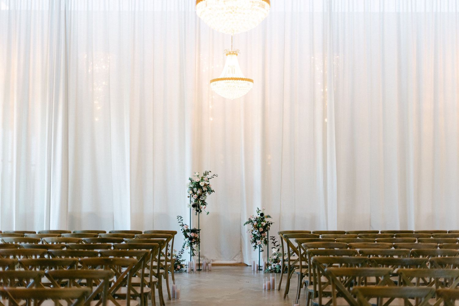 Elegant wedding ceremony venue with white draping, pendant lights, wooden chairs, and floral arrangements flanking the aisle.