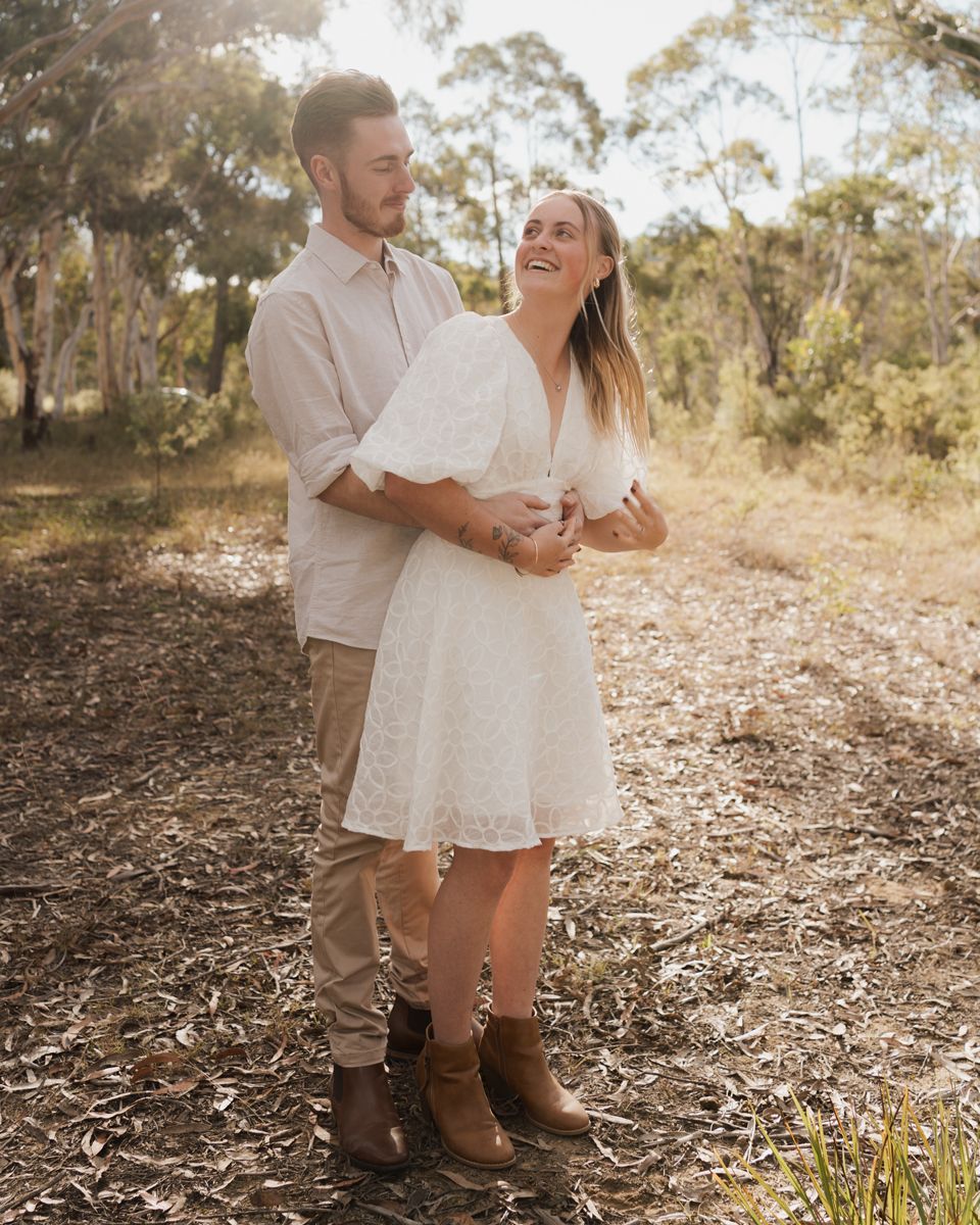Happy couple outdoors in a sunlit bushland setting, woman in white lace dress and boots, man in beige shirt engagement photoshoot rouse hill
