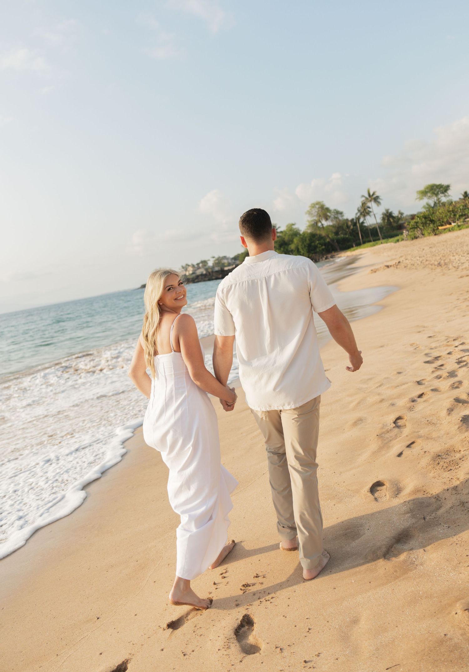 Couple holding hands at sunset by the ocean in Maui, woman in white dress, man in khaki, on lush green lawn.
