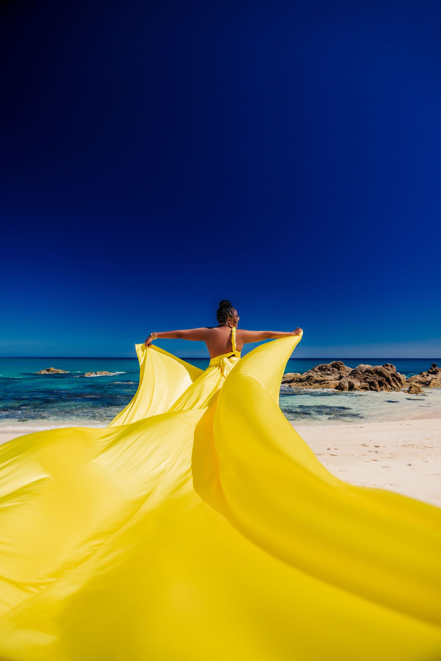 Woman spreading yellow silk fabric on white sand beach under blue sky.