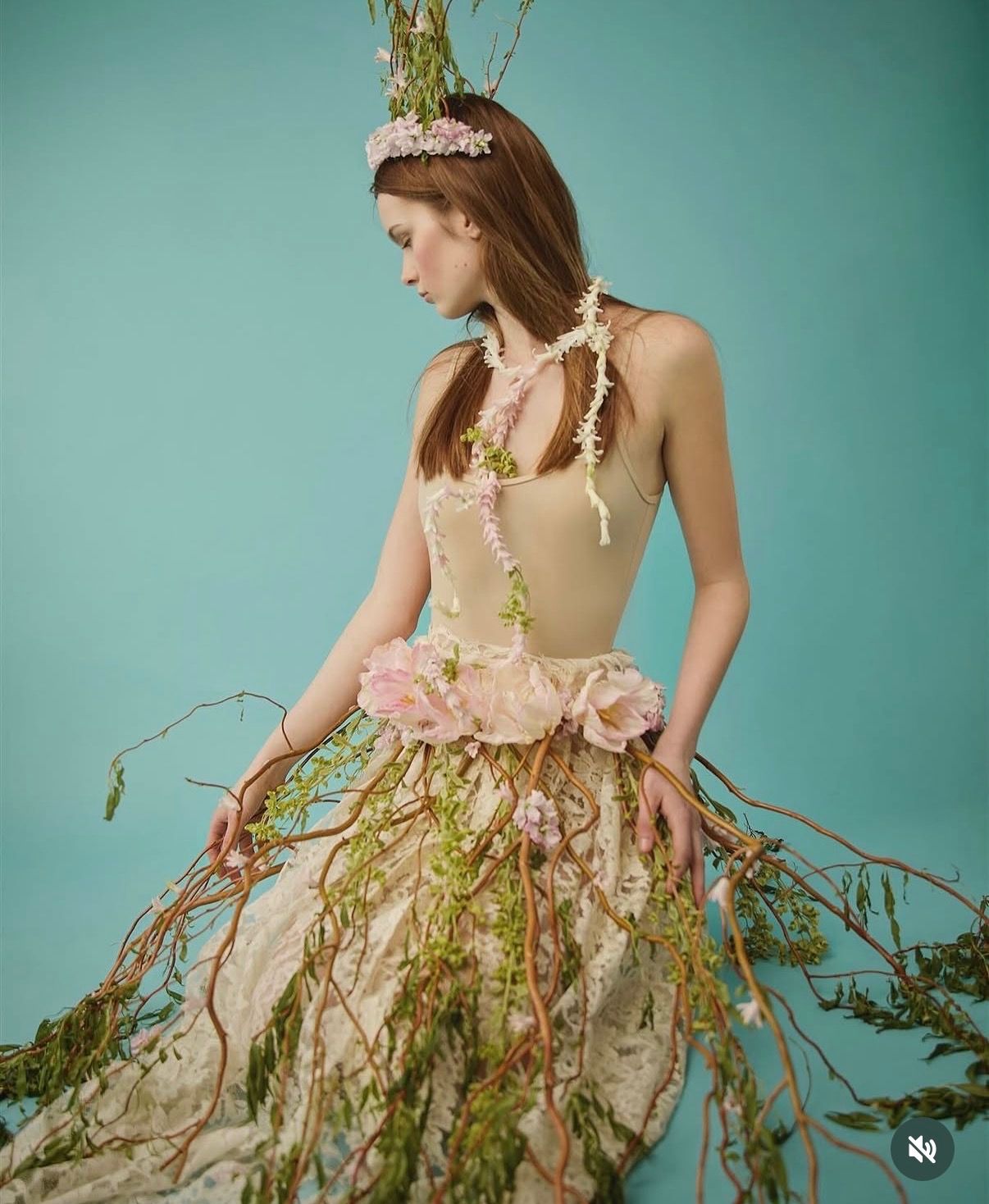 A model wears an ethereal floral gown adorned with vines, branches, and pink flowers against a teal background.