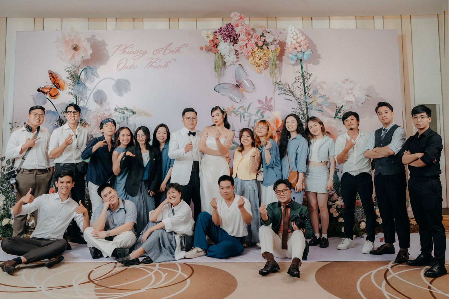 Large group of wedding guests pose and smile in front of a floral decorated reception backdrop.