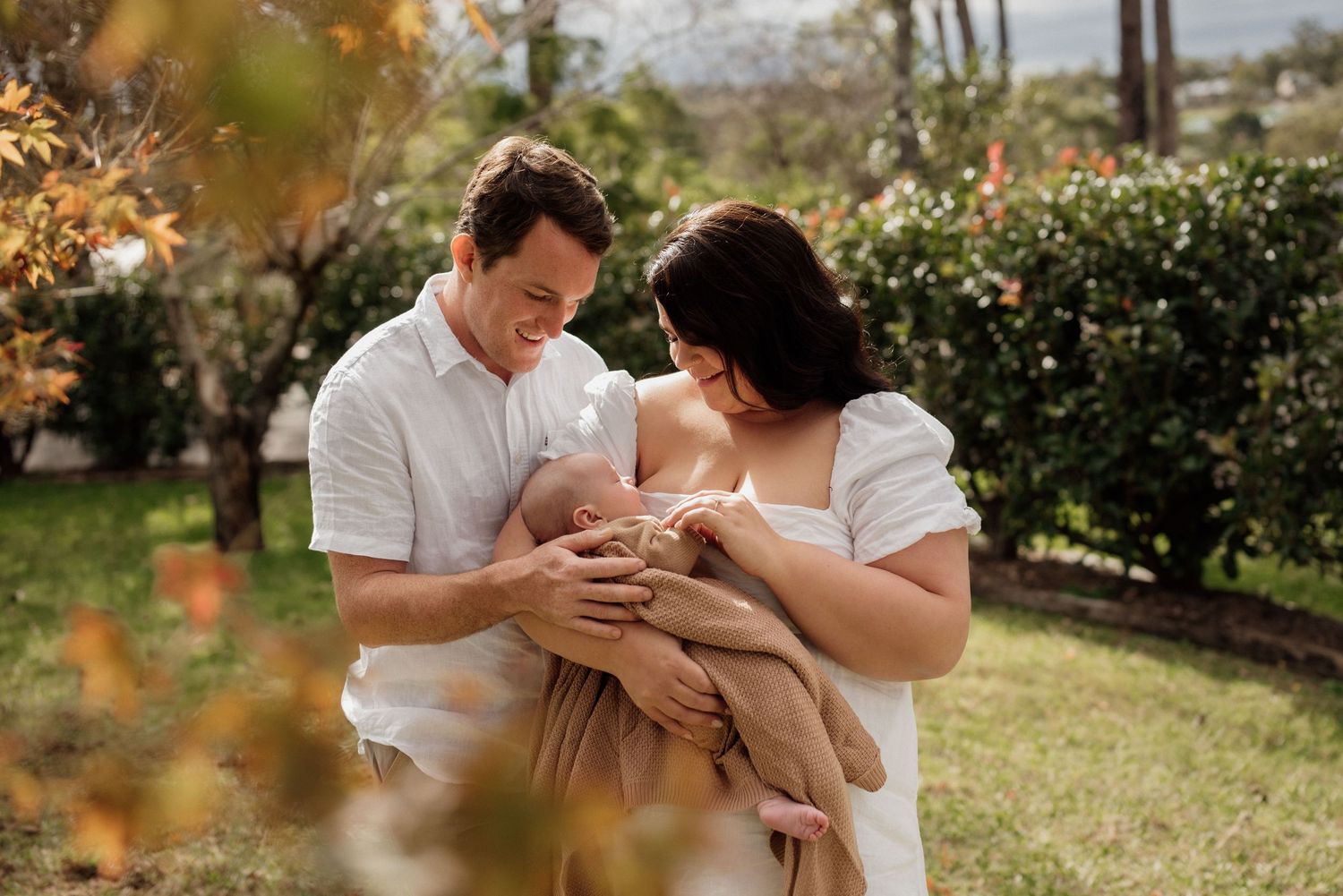 Parents lovingly kiss and cuddle their sleeping newborn baby wrapped in a white swaddle blanket.