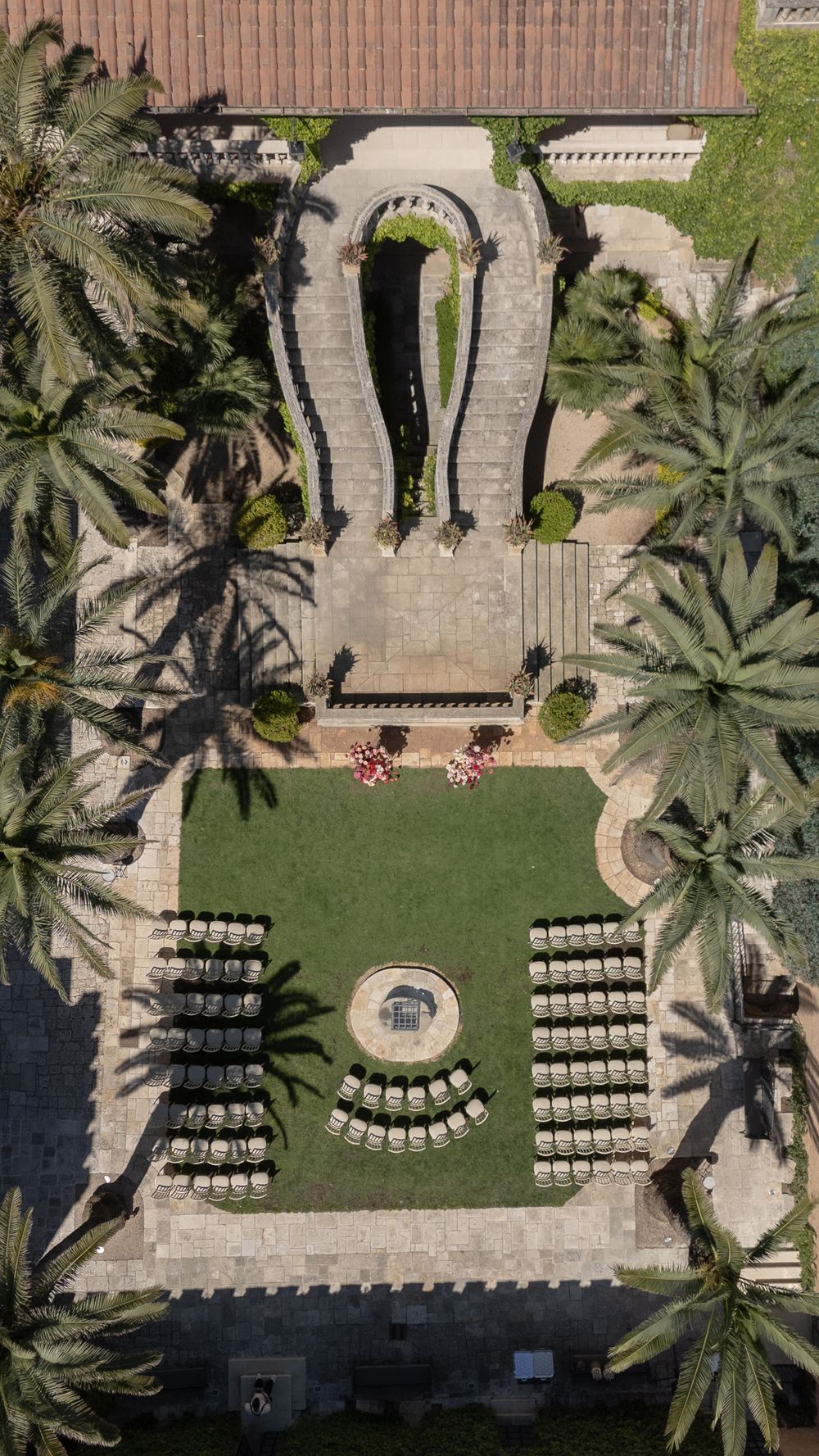 Aerial view of an elegant outdoor courtyard with symmetrical seating, a fountain, lush lawn, and palm trees surrounding a grand archway.