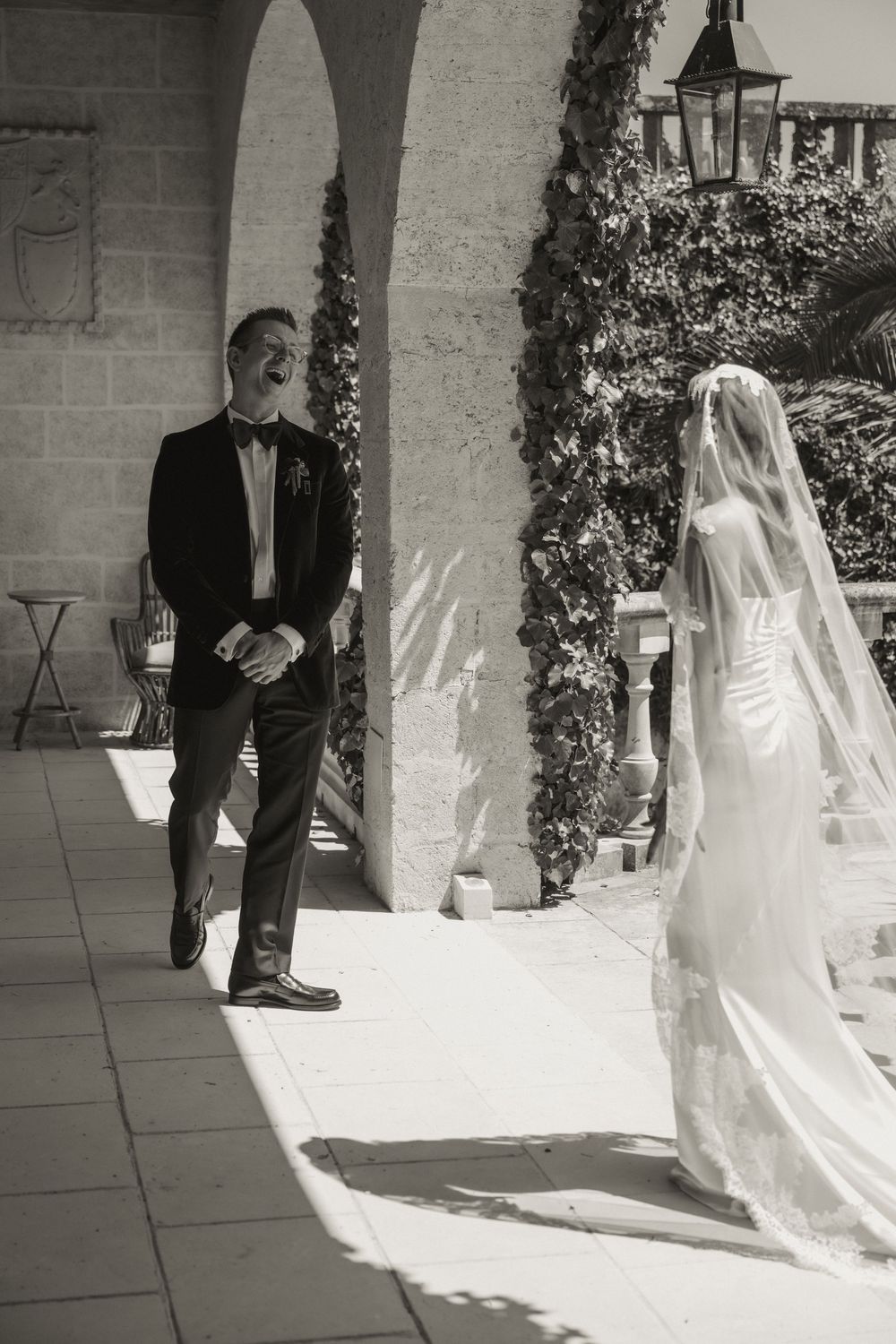 Black and white photo of groom smiling as bride in veil approaches during wedding first look at elegant venue.