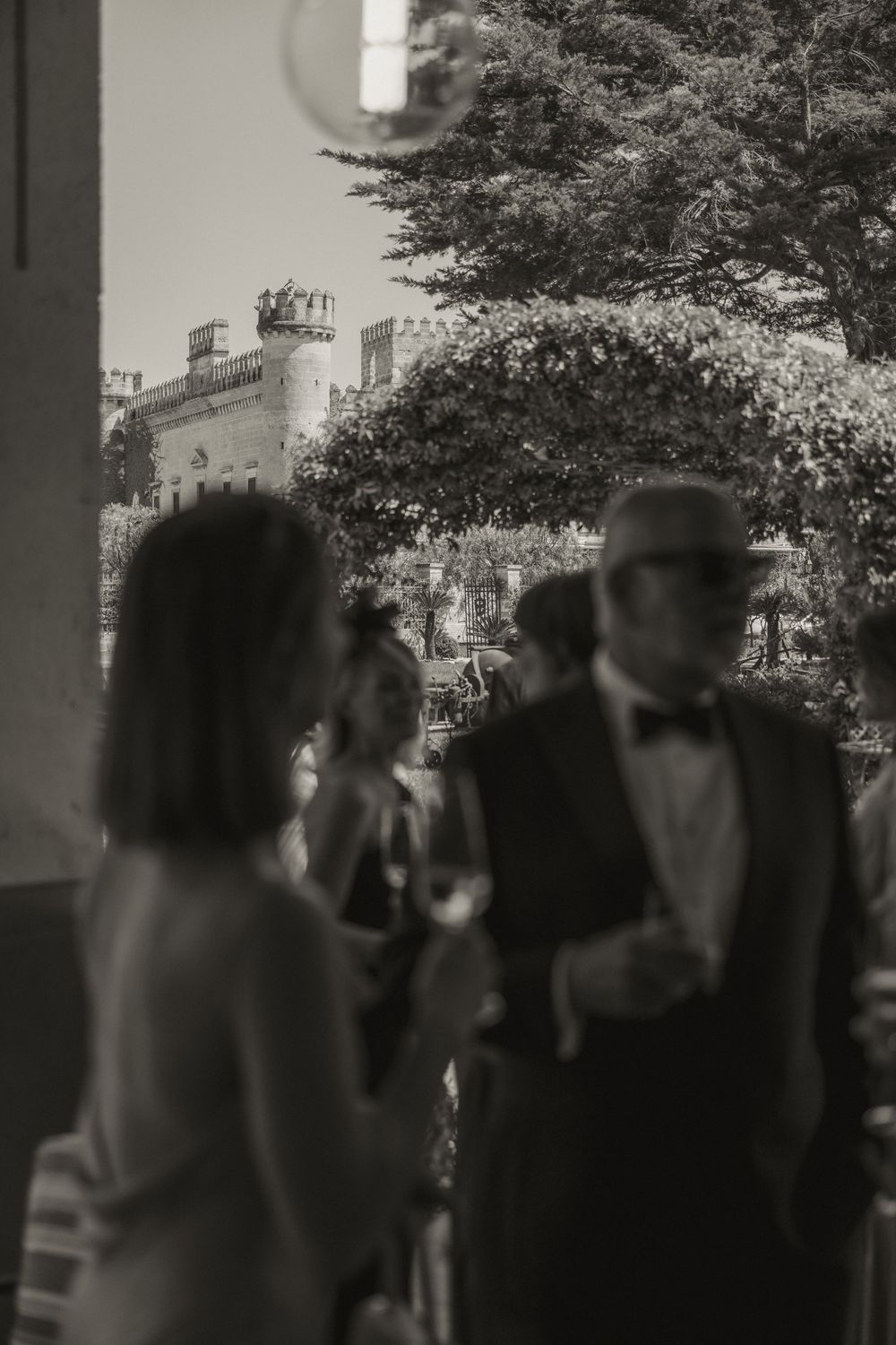 Black and white photo of elegantly dressed guests at a formal outdoor event near a historic castle.
