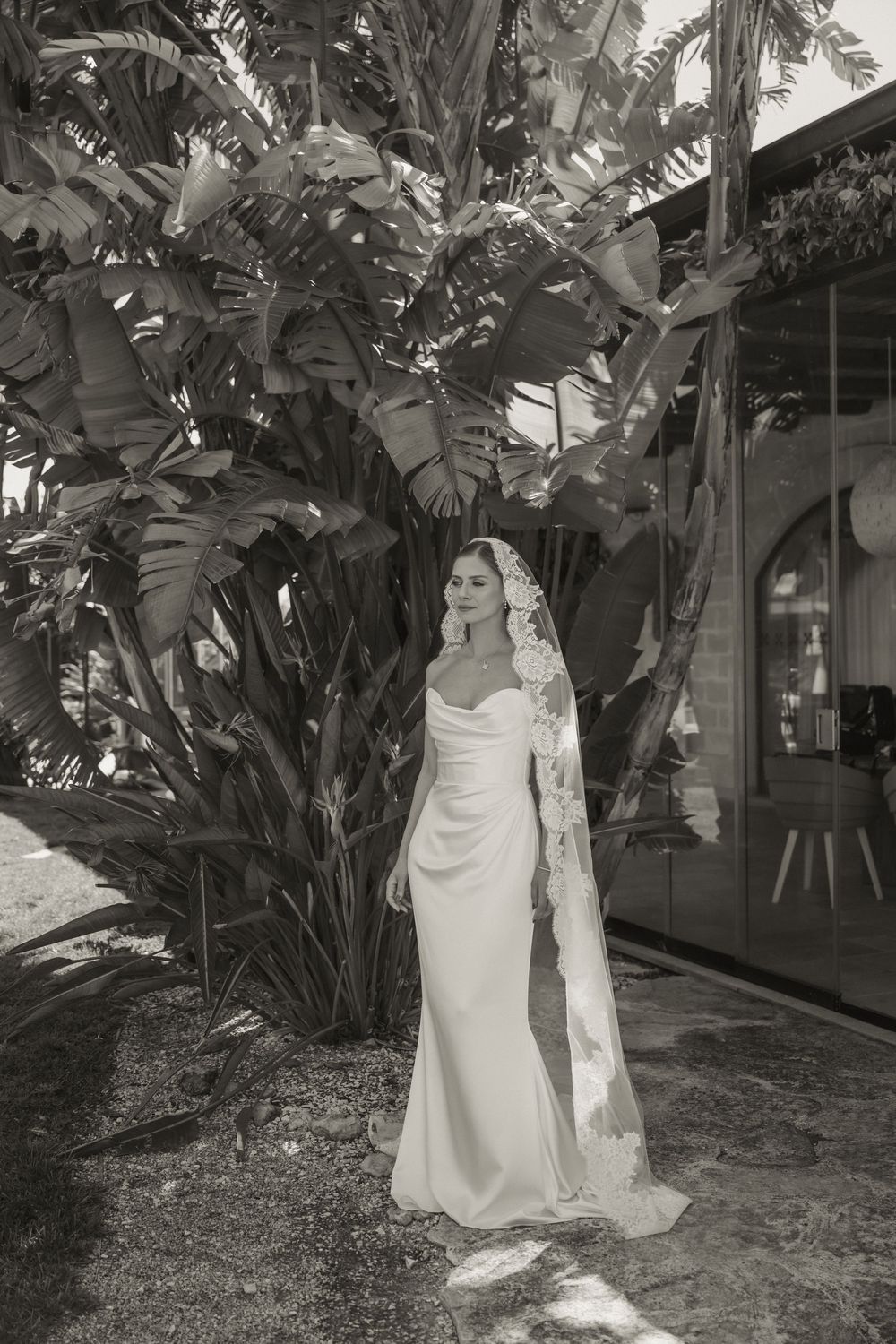 Bride in elegant white gown and lace veil stands before tropical foliage in black and white wedding portrait.