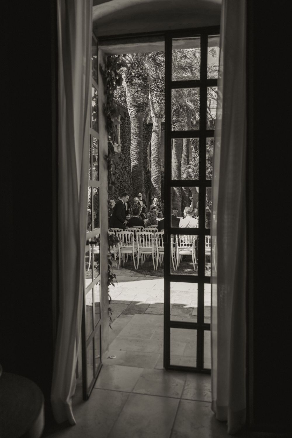 Black and white view through open doors of an outdoor wedding reception with white chairs among palm trees.