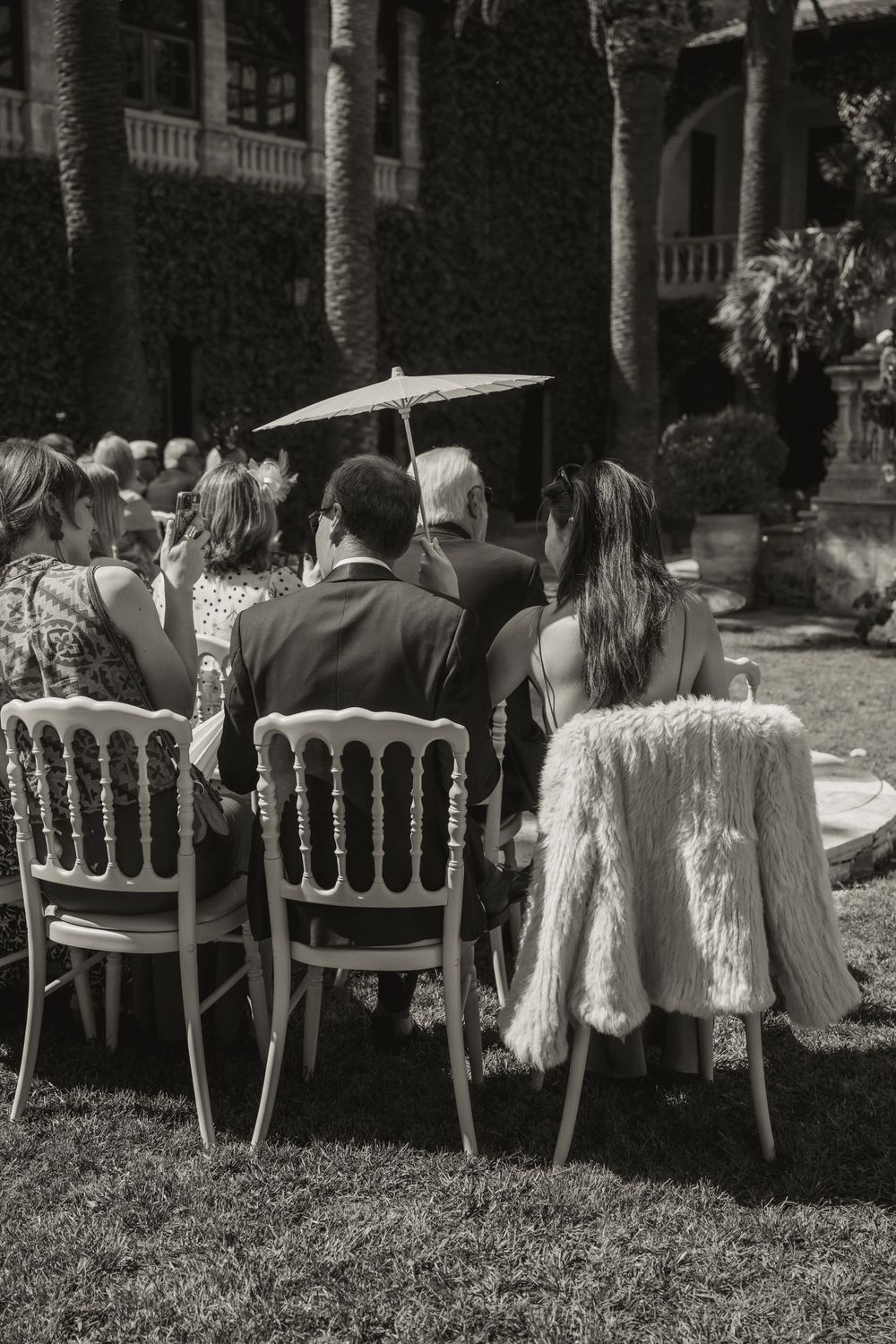 Black and white photo of wedding guests seated on elegant chairs in a garden ceremony outdoors.