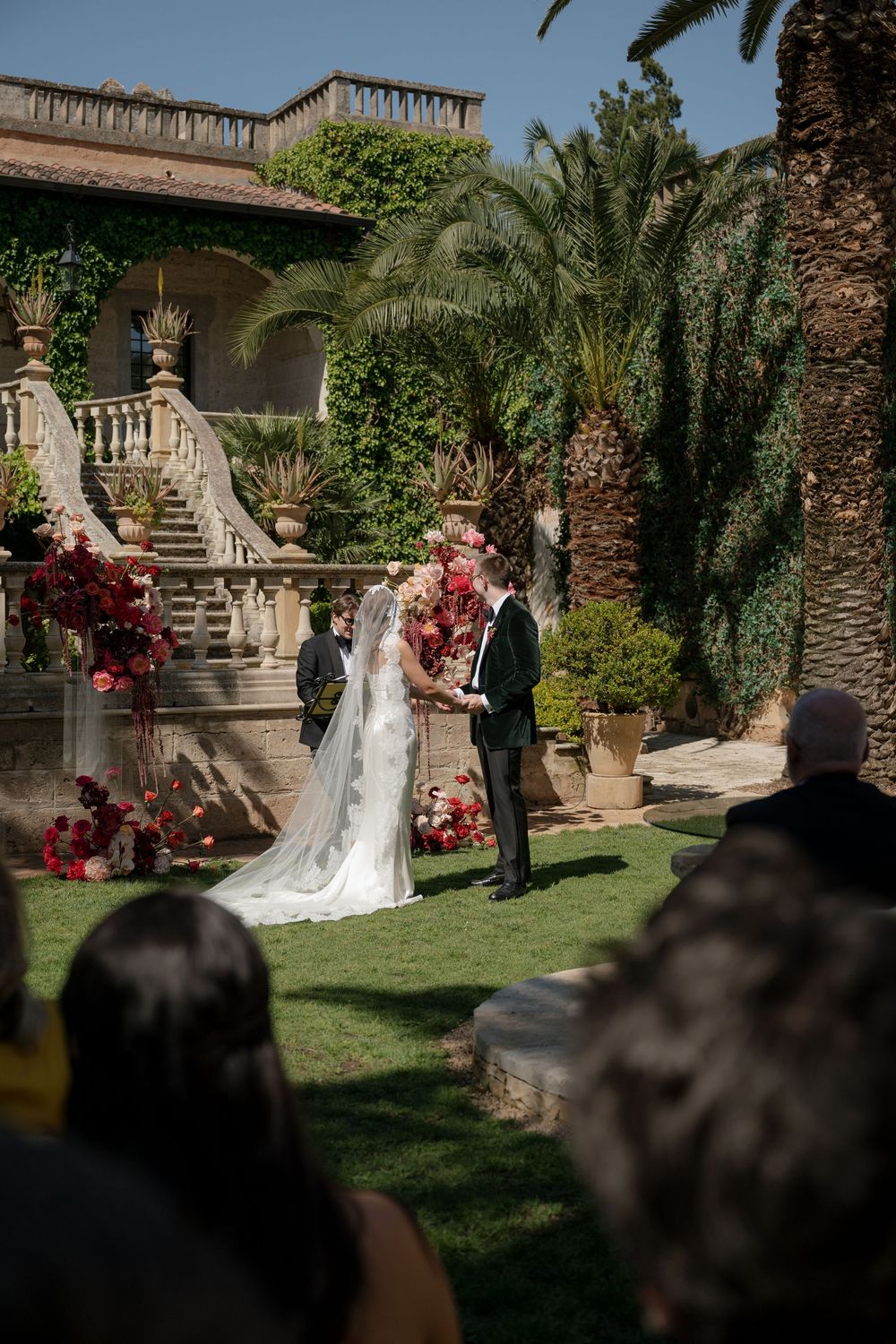 Outdoor wedding ceremony on lush lawn with bride and groom at floral arch, elegant villa staircase and palm trees backdrop.