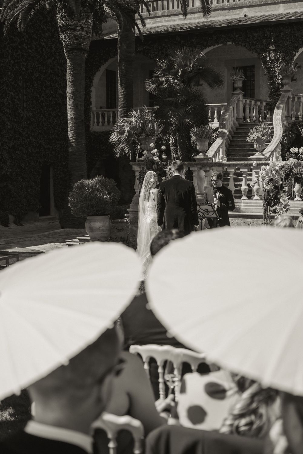 Black and white wedding photo of bride and groom at elegant villa with guests holding white parasols in foreground.